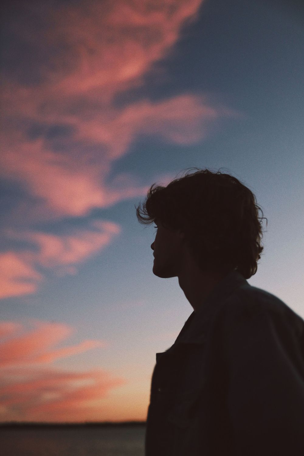 A silhouetted figure with curly hair stands against a dramatic sunset sky with pink clouds.