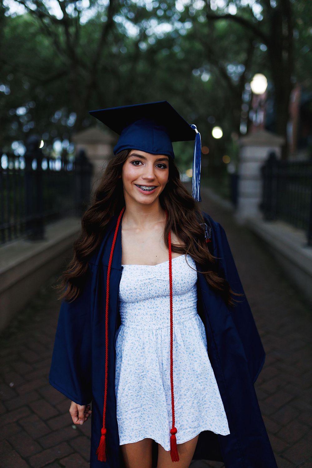 Graduate in navy cap and gown with white top poses outdoors on a tree-lined path at dusk.