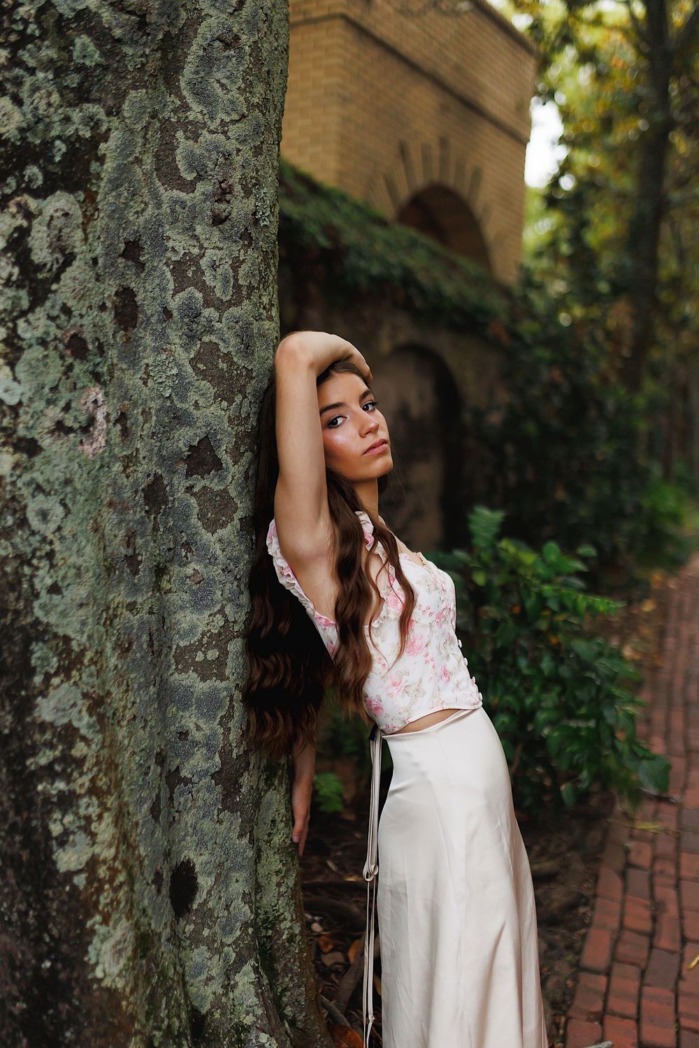 A woman in a white dress poses artistically against a tree trunk in a garden setting with brick pathways and arches.