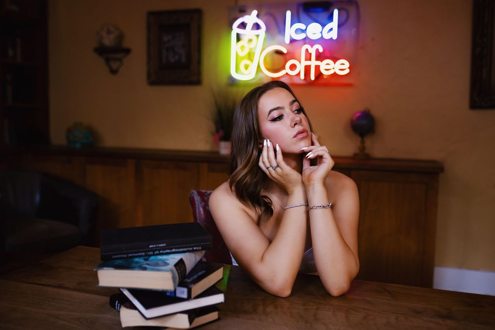 A person sits at a wooden table with books while daydreaming under a glowing neon 'Iced Coffee' sign.