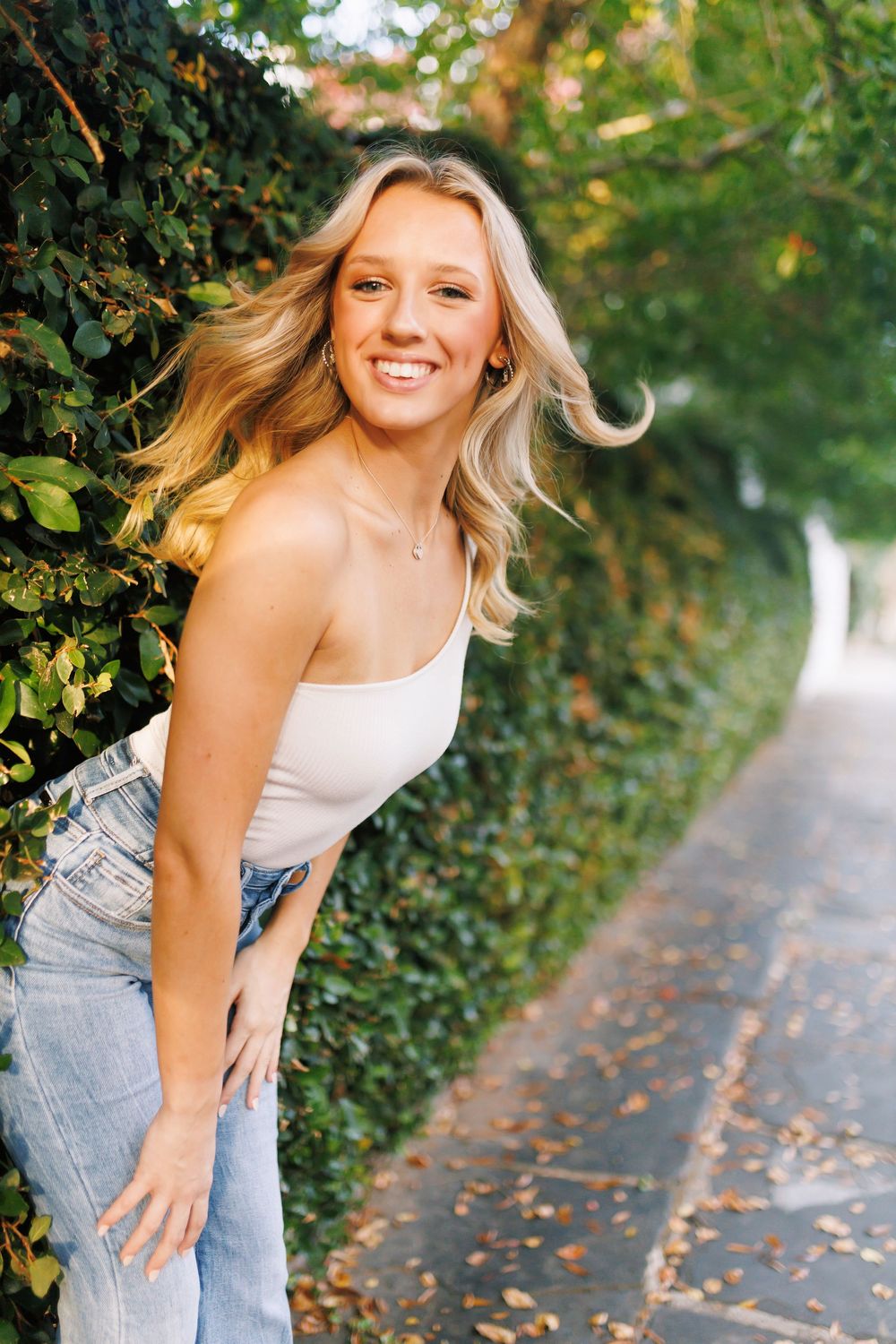 A person in jeans and white top poses playfully against a green hedge along a path with scattered autumn leaves.