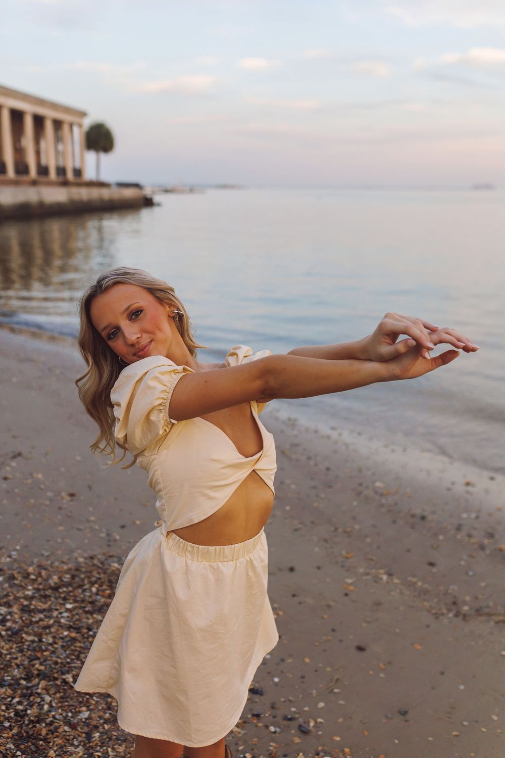 Woman in cream dress dances gracefully on beach at sunset near pier.