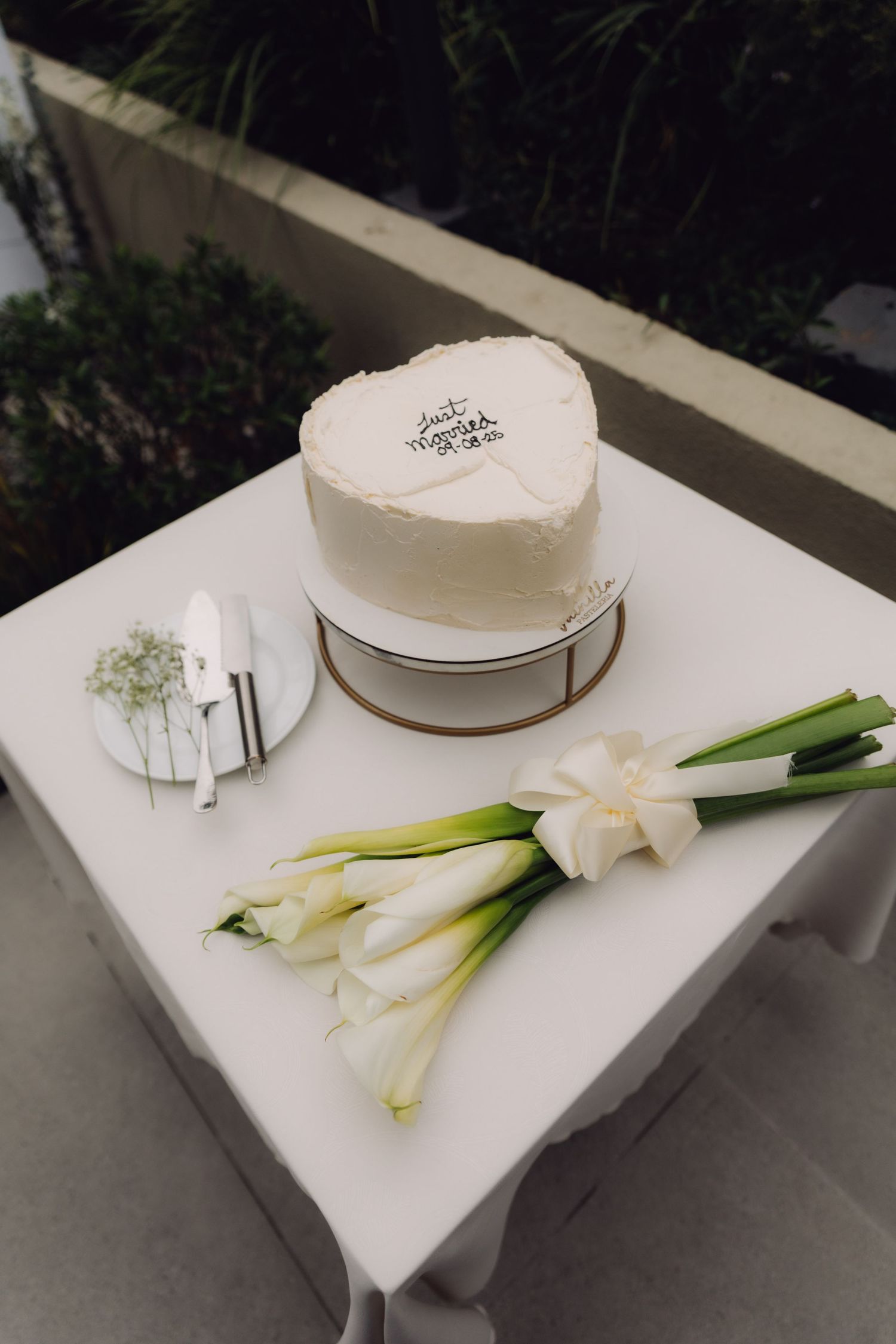 Heart-shaped Just Married wedding cake beside white calla lily bouquet on table.