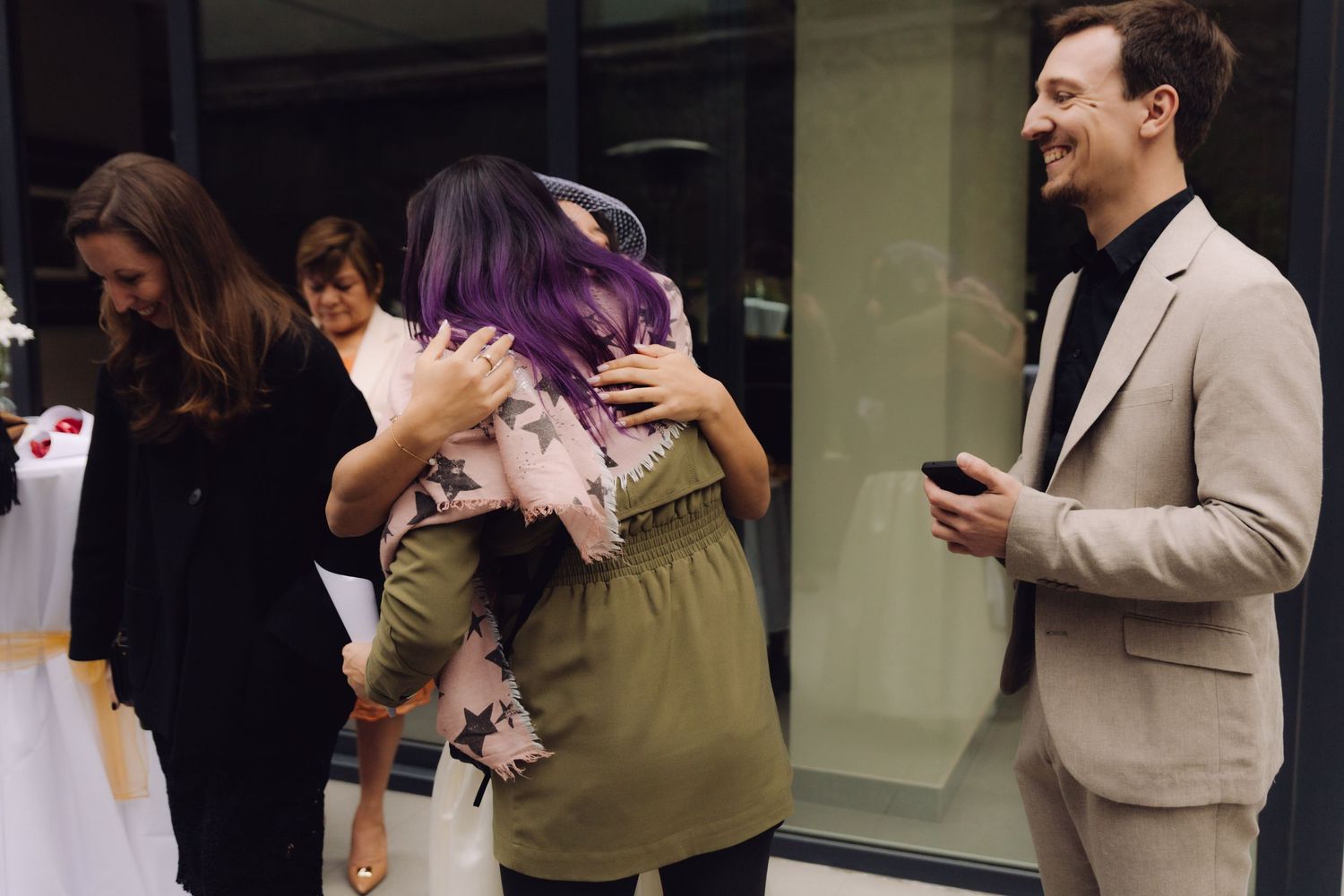 Wedding guests hugging and celebrating at an outdoor reception with a smiling man in a beige suit.