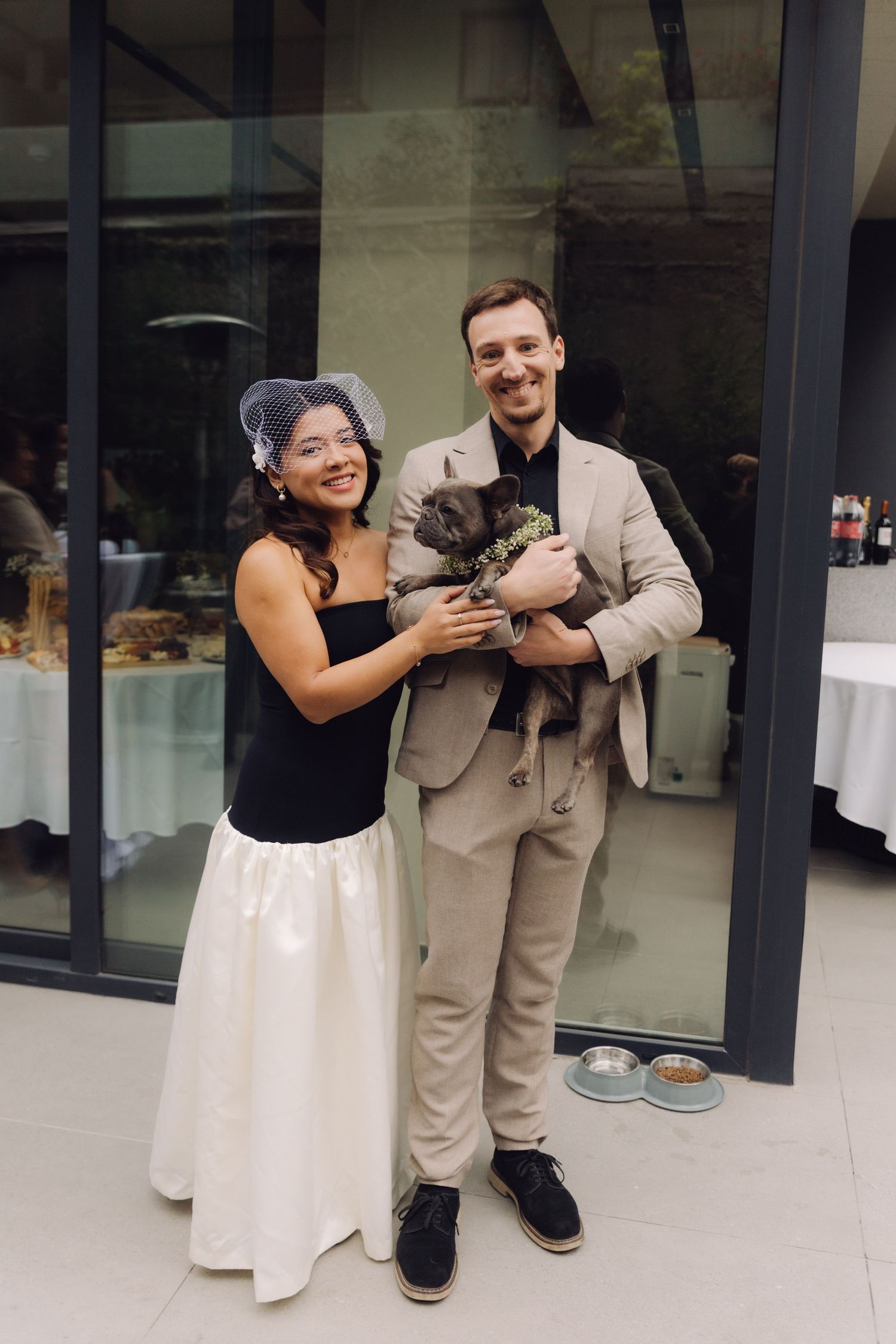 Smiling newlyweds in elegant attire holding their small dog dressed up at an outdoor wedding celebration.