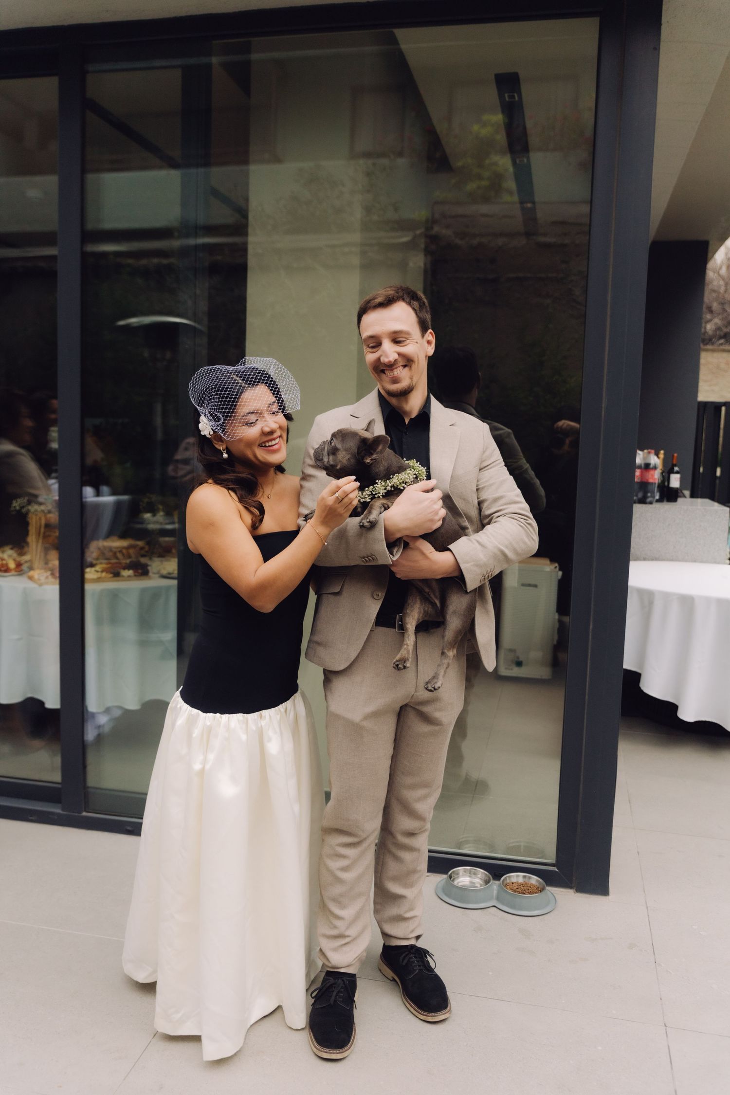 Bride in black top and white skirt with groom in beige suit holding a small dog at wedding reception.