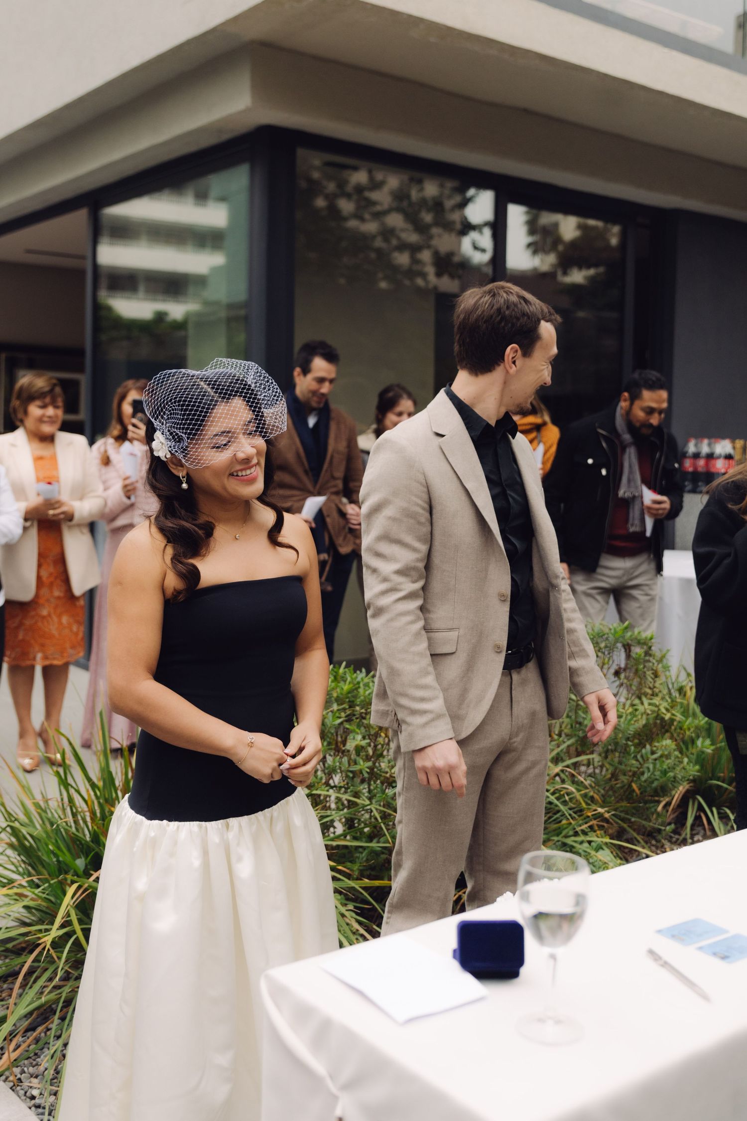 Bride in black strapless top and birdcage veil smiling at outdoor wedding ceremony with guests.