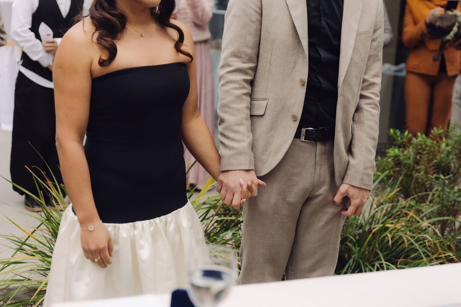 Newlyweds holding hands at wedding ceremony, bride in black strapless gown and white skirt.