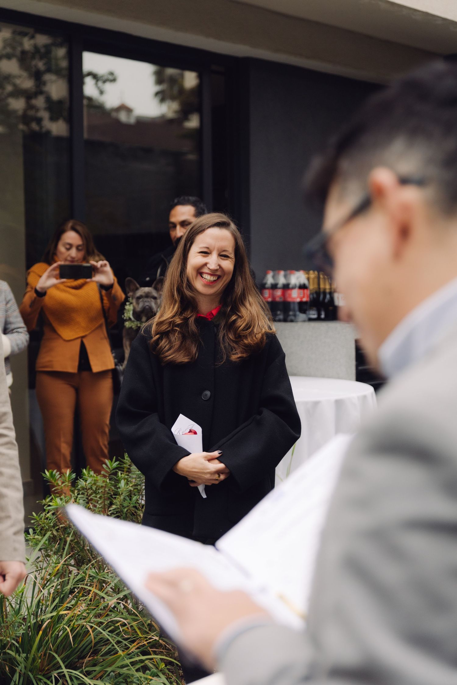 Smiling female officiant in black robe conducting an outdoor wedding ceremony for guests.