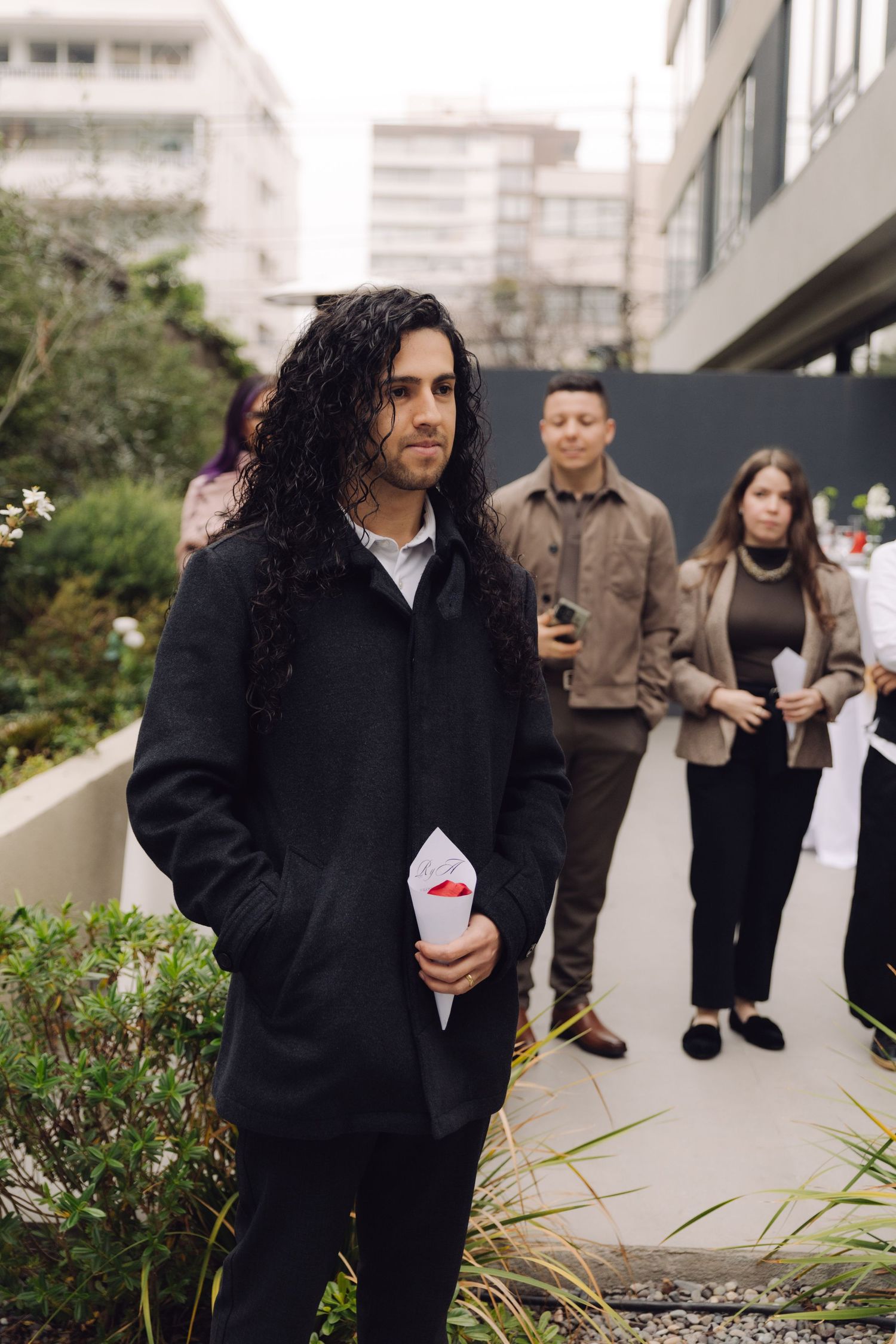Wedding guest with long curly hair in black coat holding ceremony program at outdoor wedding.