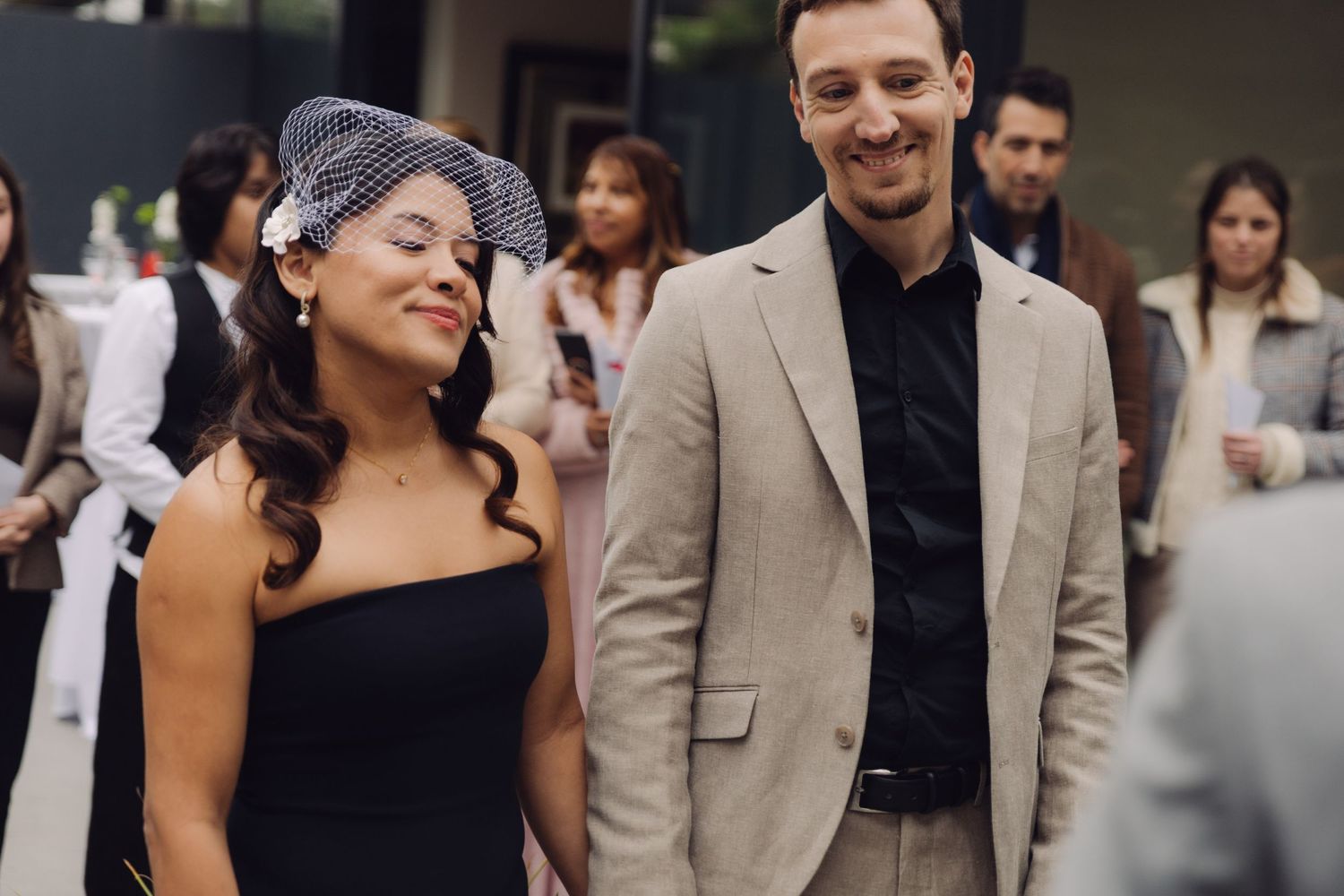 Bride wearing birdcage veil and black strapless dress walking with groom in beige suit at wedding.