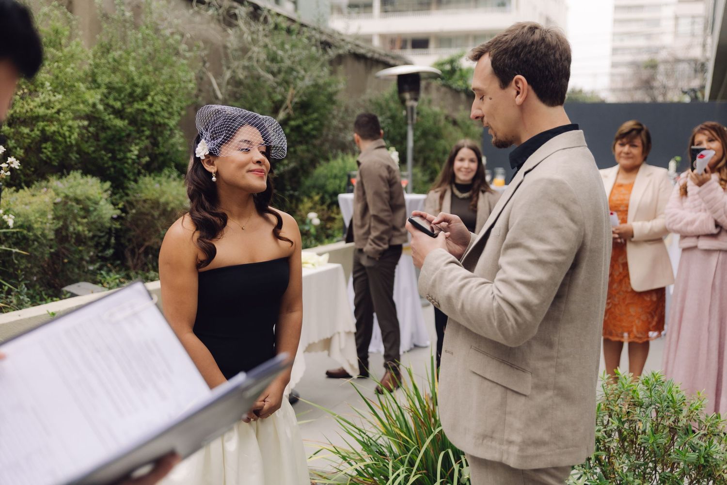 Bride in birdcage veil smiles during outdoor wedding ceremony with guests.