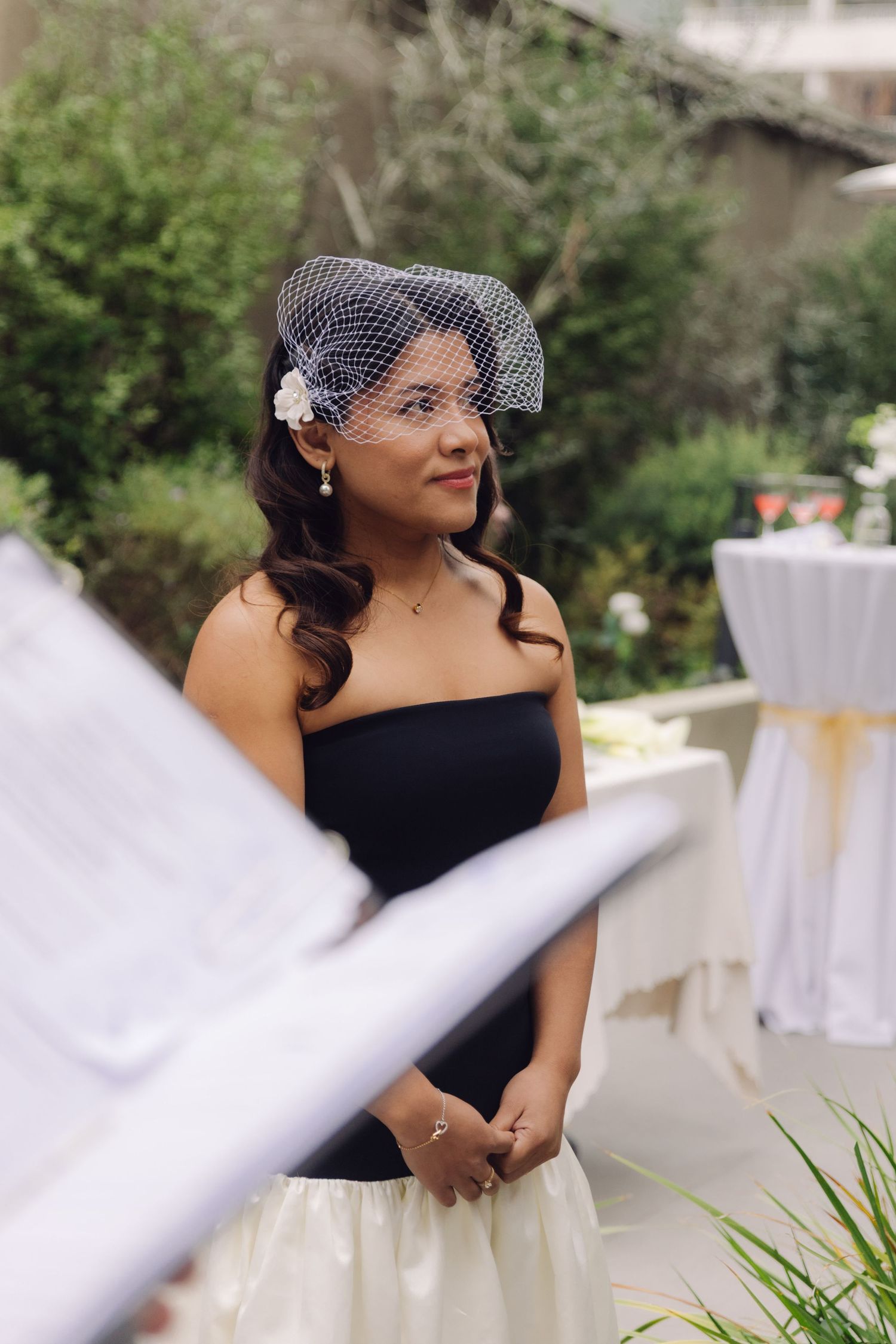 Bride in black strapless gown and birdcage veil with white flower stands outdoors at wedding ceremony.