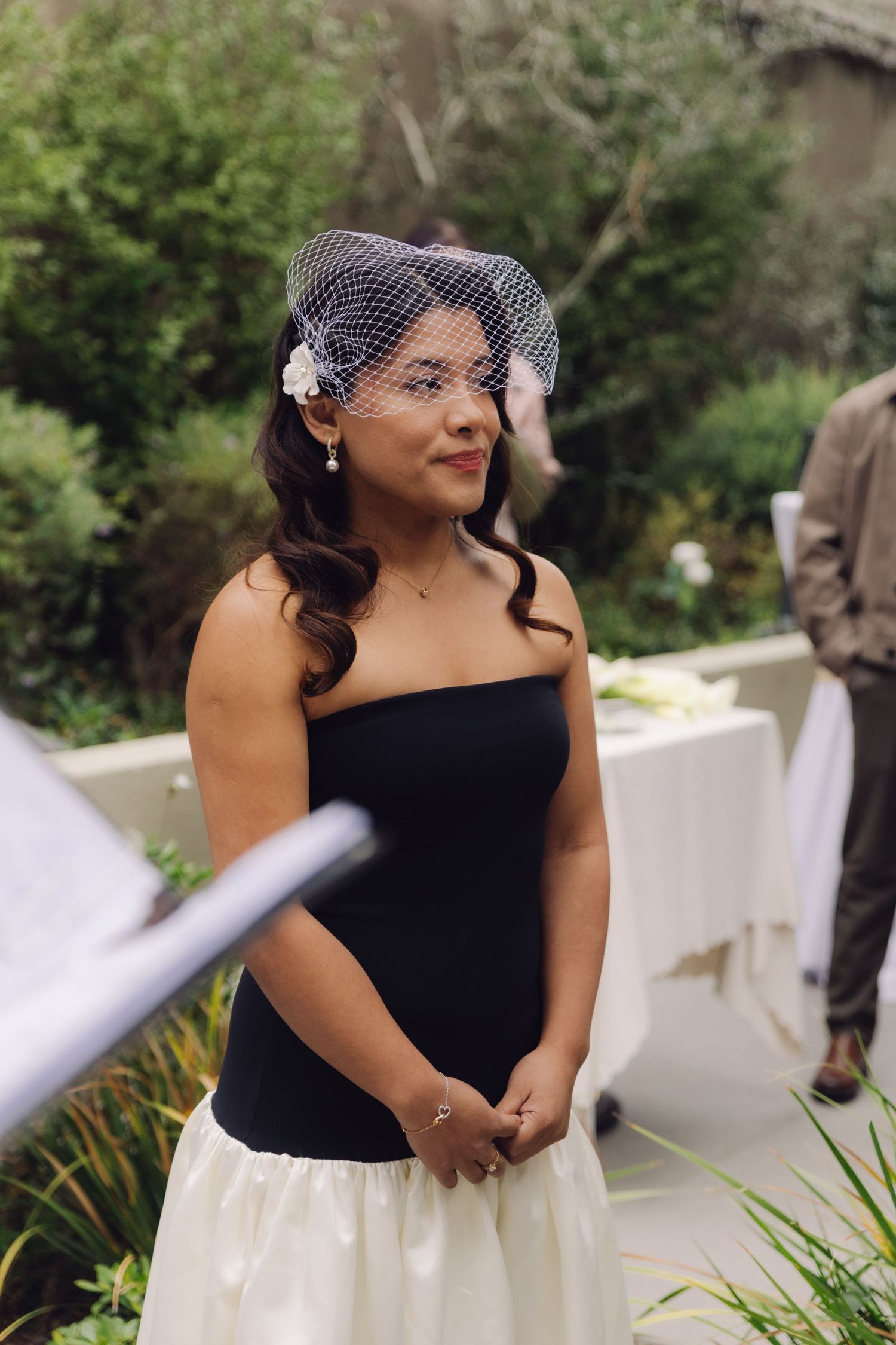 Beautiful bride in black strapless gown and white birdcage veil smiling at outdoor wedding ceremony.