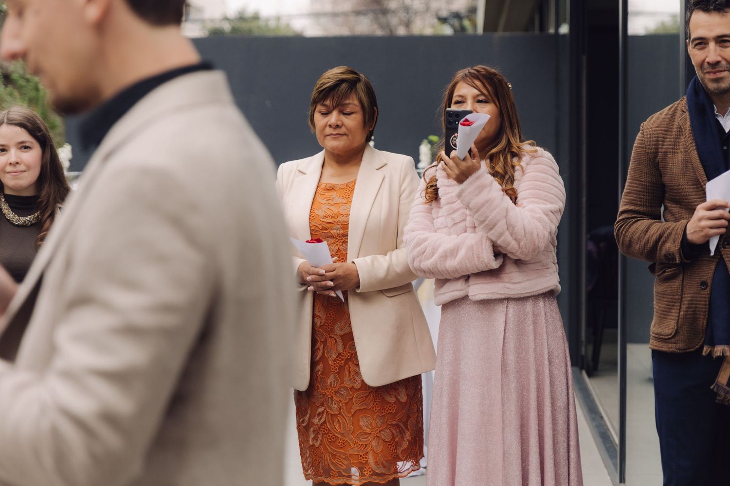 Wedding guests in pink and orange dresses watch outdoor ceremony, one holding phone to record the moment.