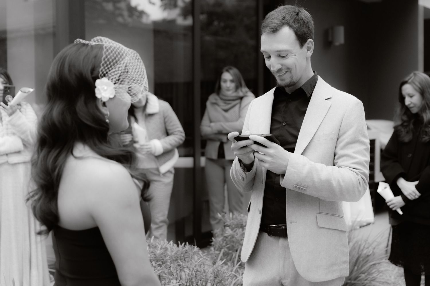 Black and white photo of groom in suit reading vows to bride wearing birdcage veil at wedding ceremony.