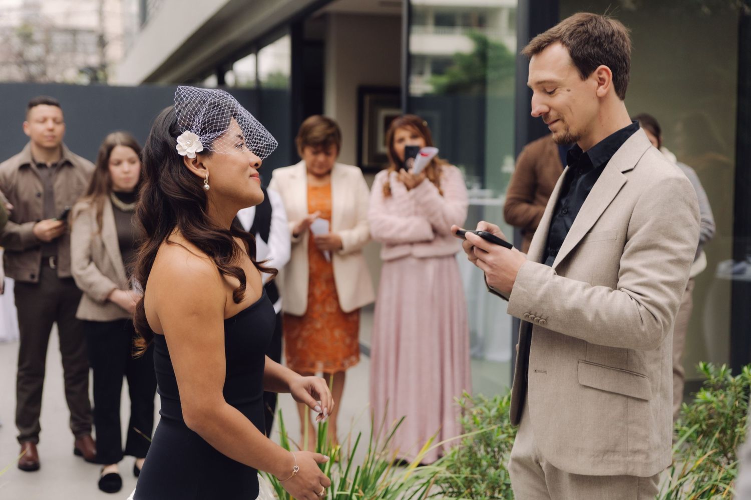 Bride in black dress and birdcage veil smiles at groom in tan suit during outdoor wedding ceremony.