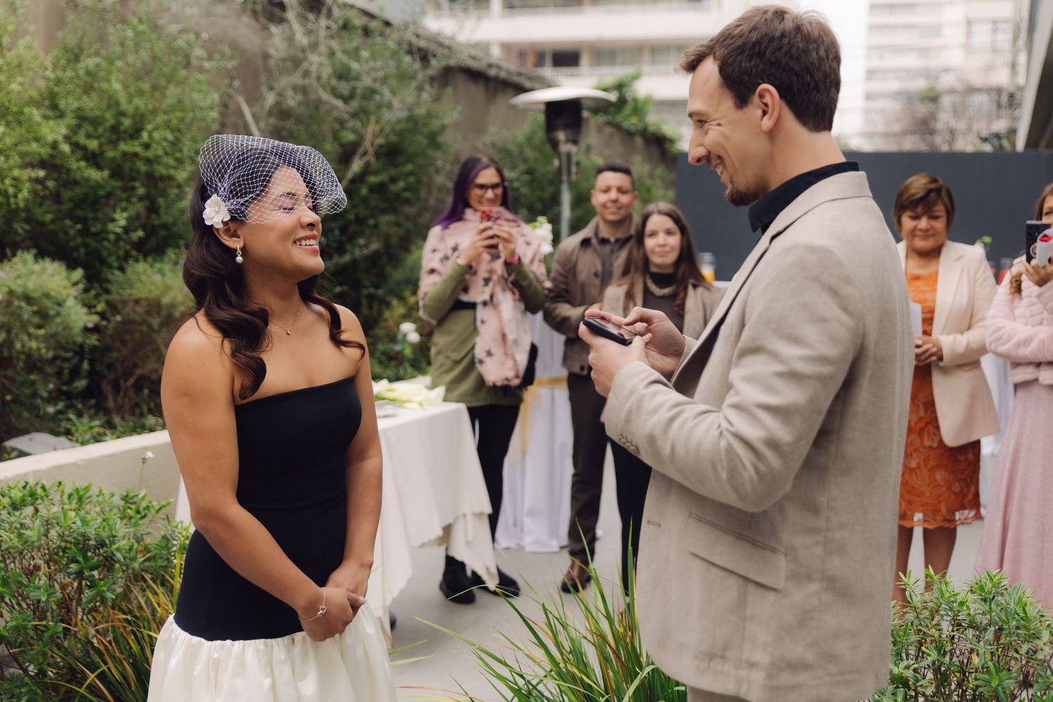 Bride laughs joyfully as groom reads vows during intimate outdoor wedding ceremony with guests watching.