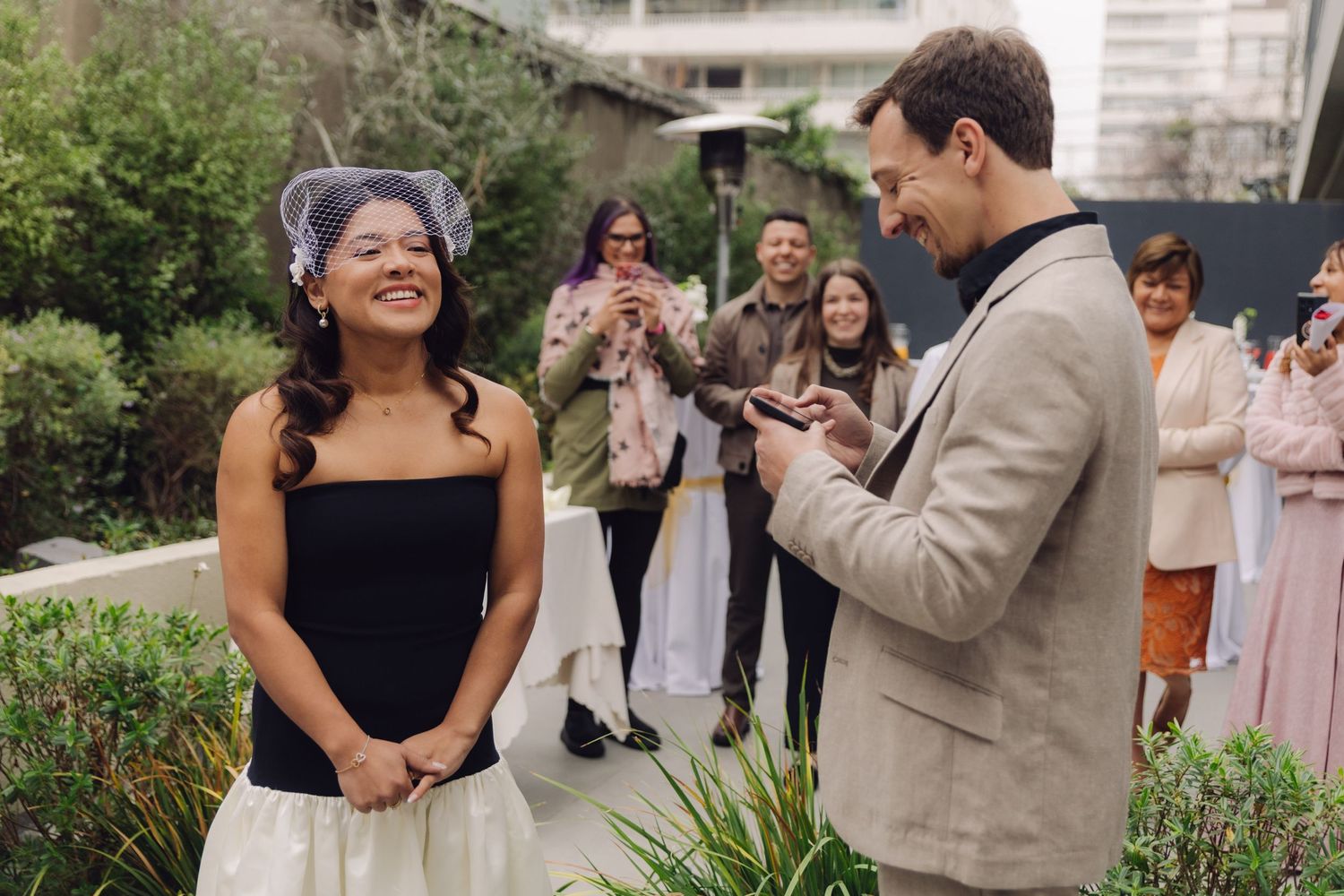Smiling bride in black strapless dress and birdcage veil listens to groom reading vows at outdoor wedding.