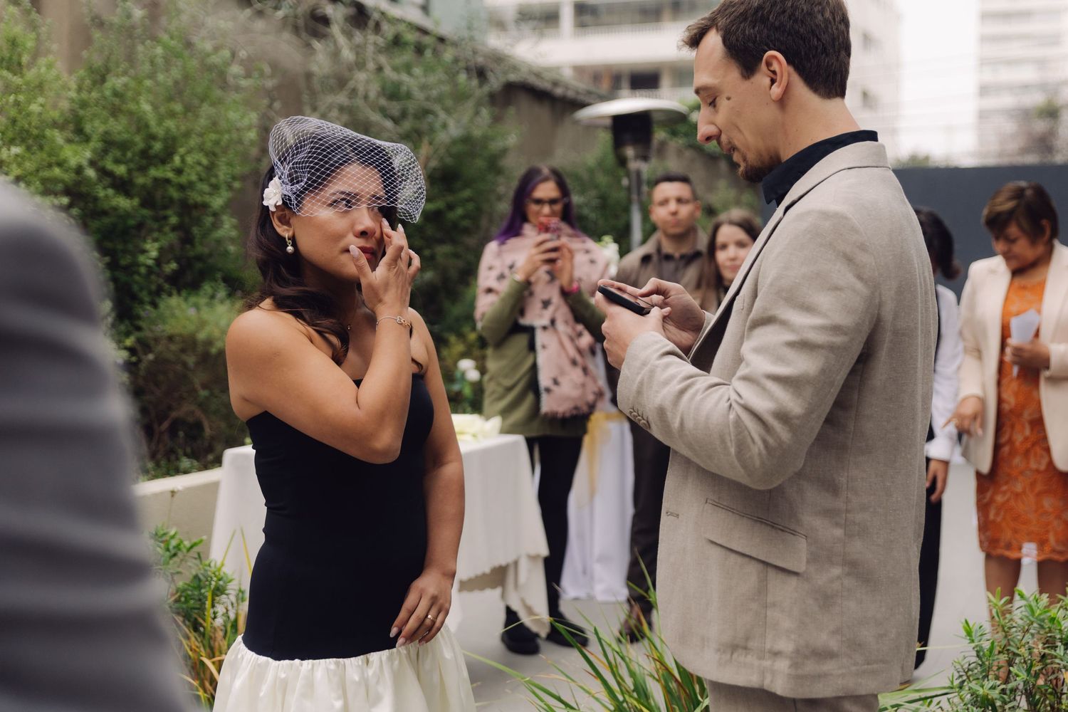 Emotional bride wipes tear as groom reads vows during outdoor wedding ceremony with guests in background.