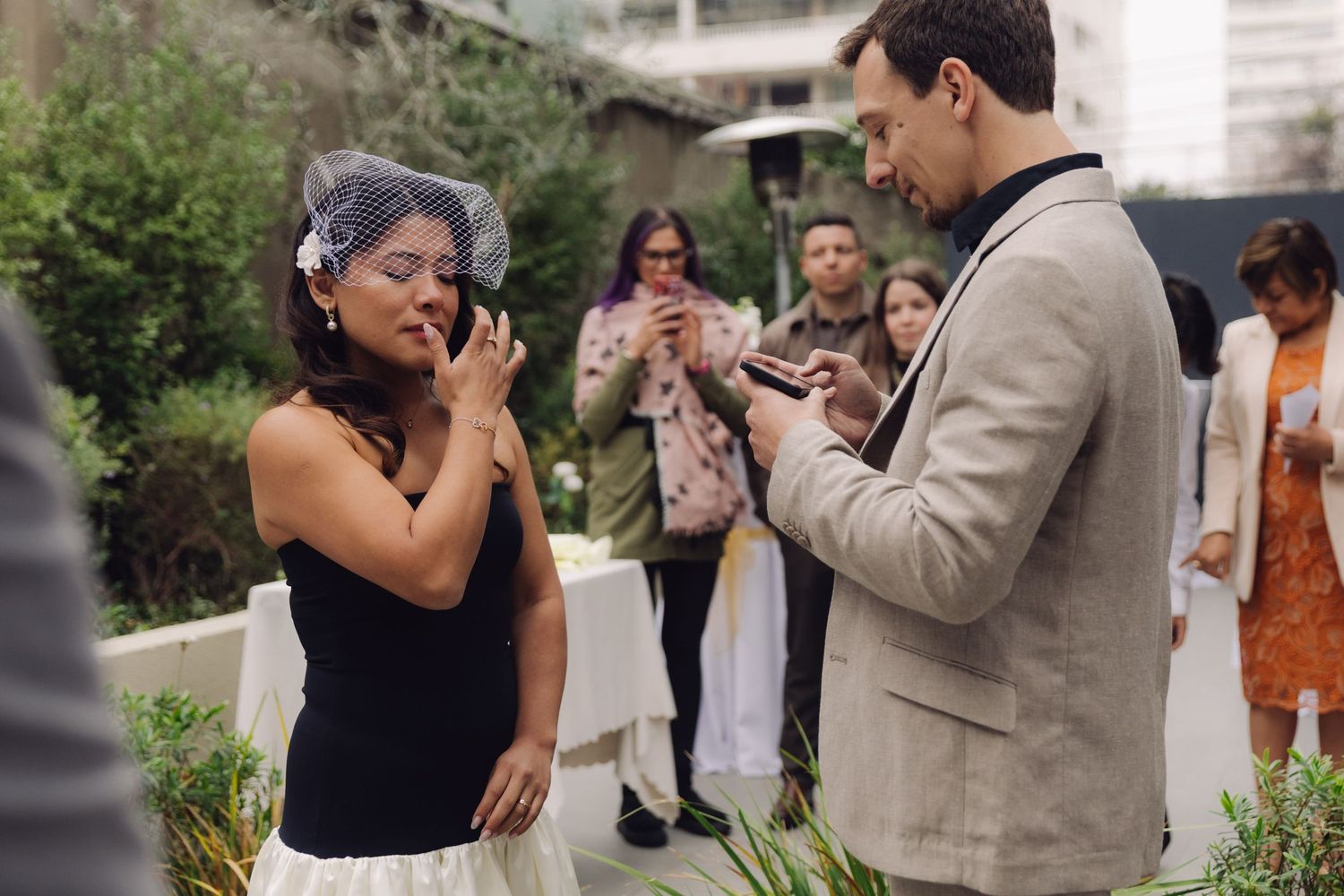 Bride touches face emotionally while groom reads from phone during outdoor wedding ceremony with guests.