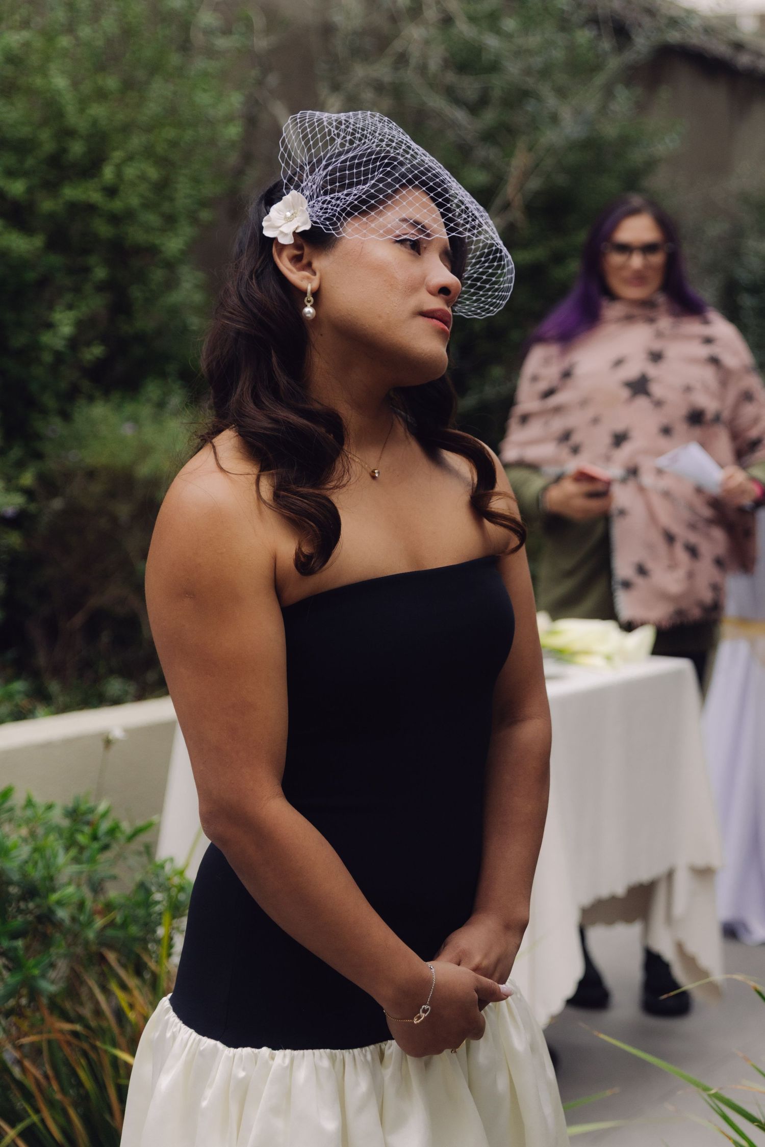 Bride in black strapless dress and birdcage veil stands attentively during outdoor wedding ceremony.