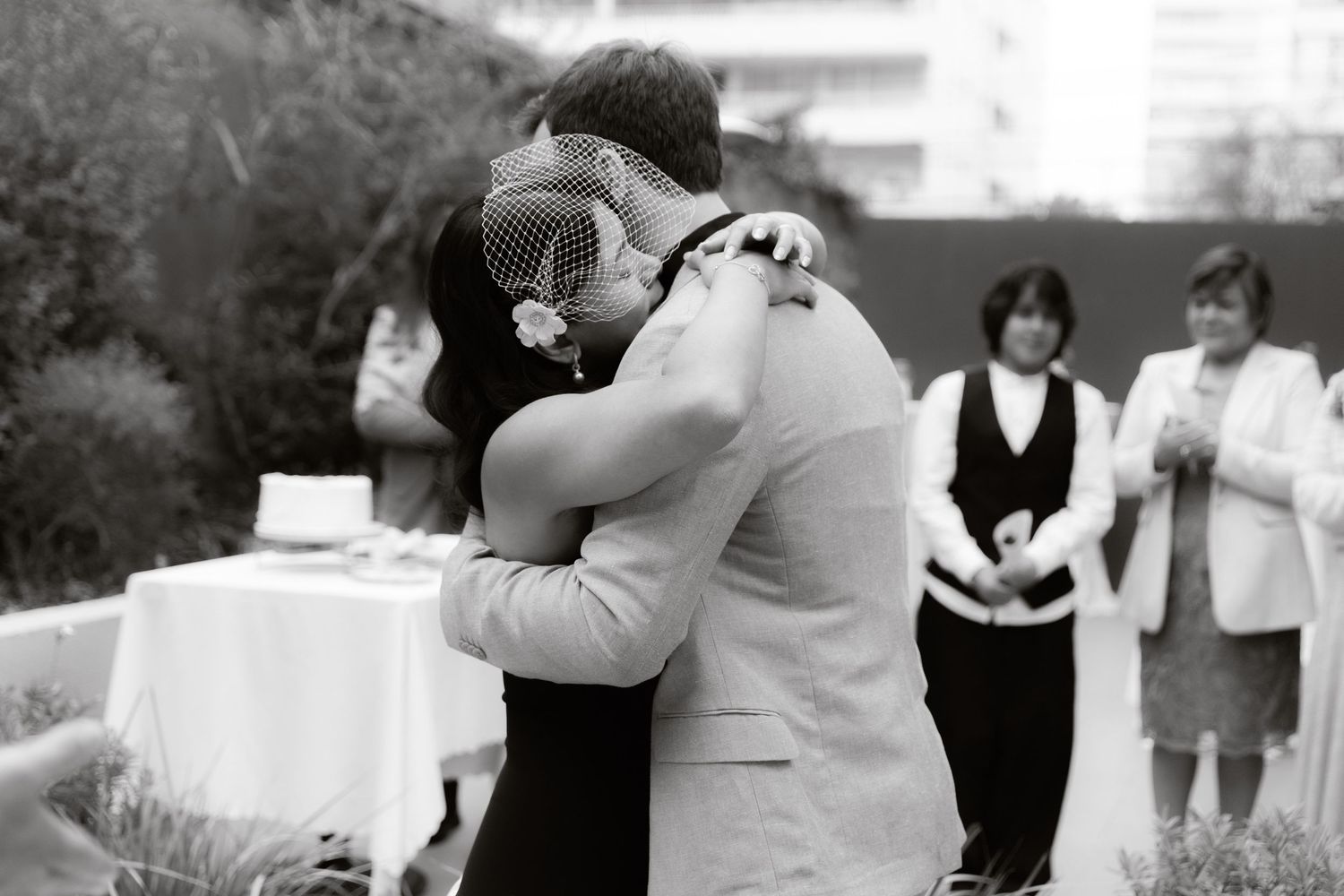 Black and white photo of newlyweds sharing first embrace after wedding ceremony with guests in background.