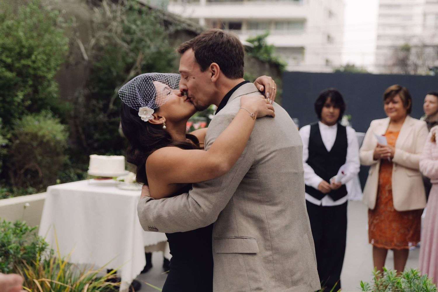 Couple sharing first kiss at outdoor wedding ceremony with guests watching in background.