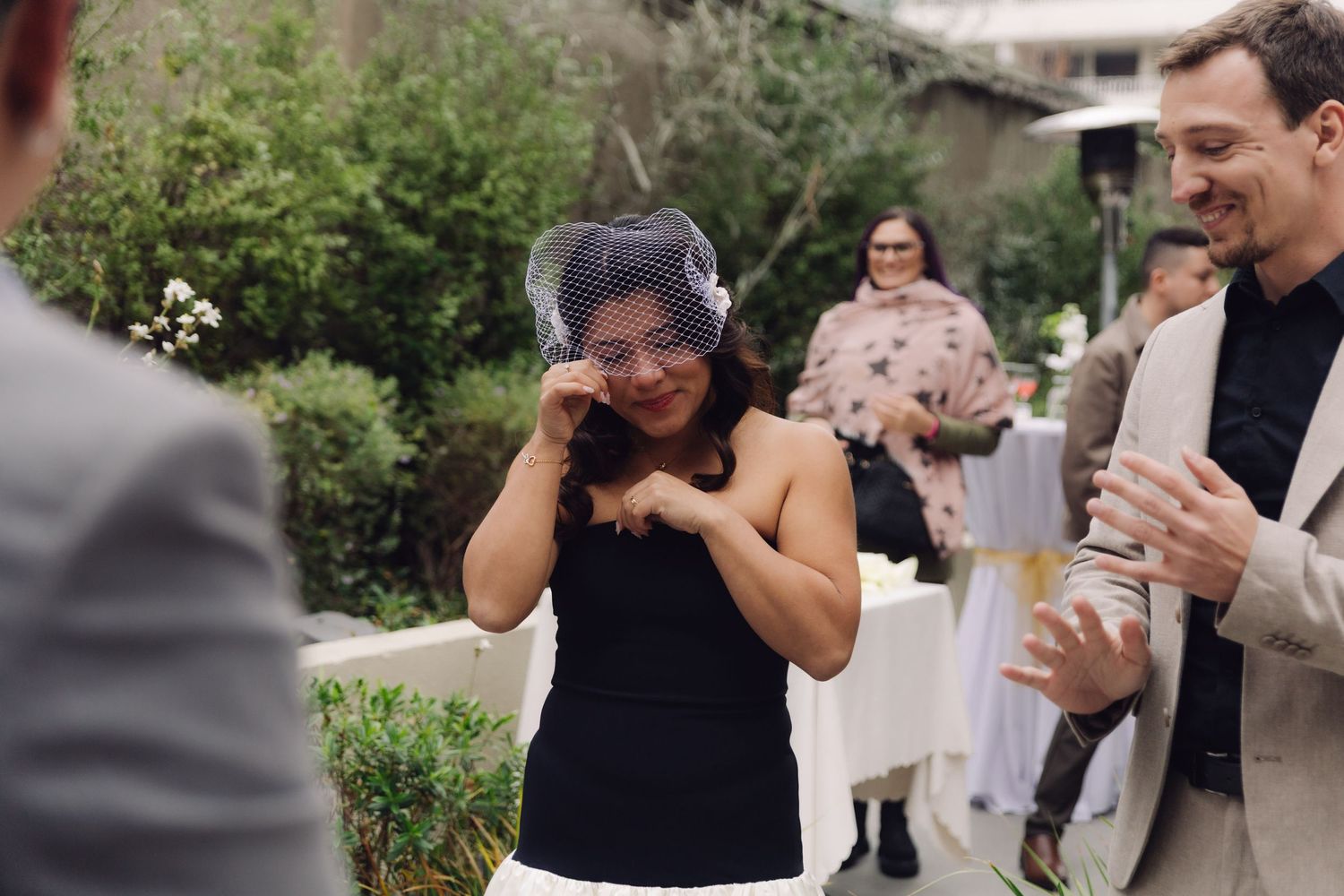 Emotional bride wiping tears wearing birdcage veil at outdoor wedding ceremony.