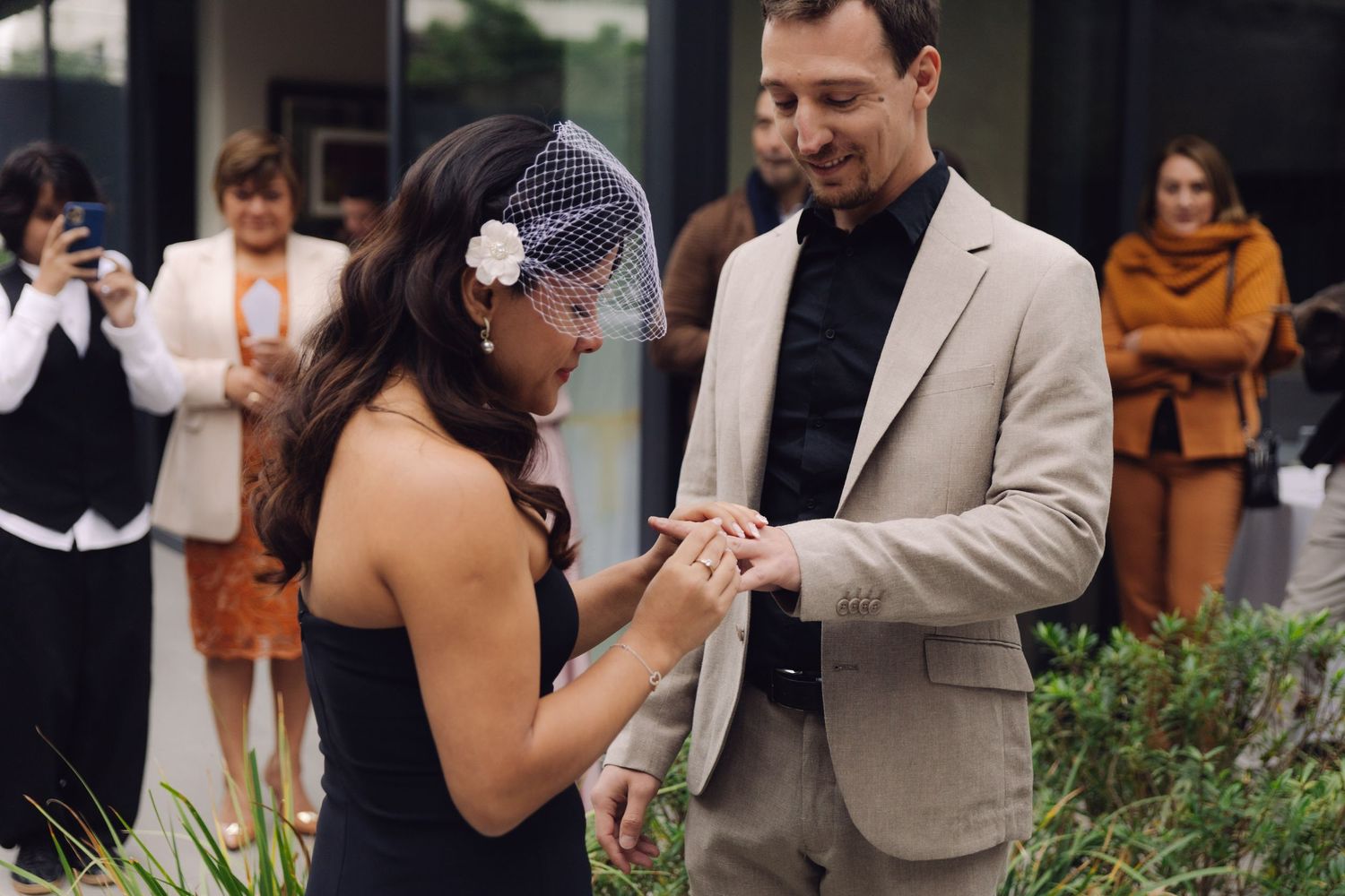 Bride placing wedding ring on grooms finger during intimate outdoor ceremony with guests.