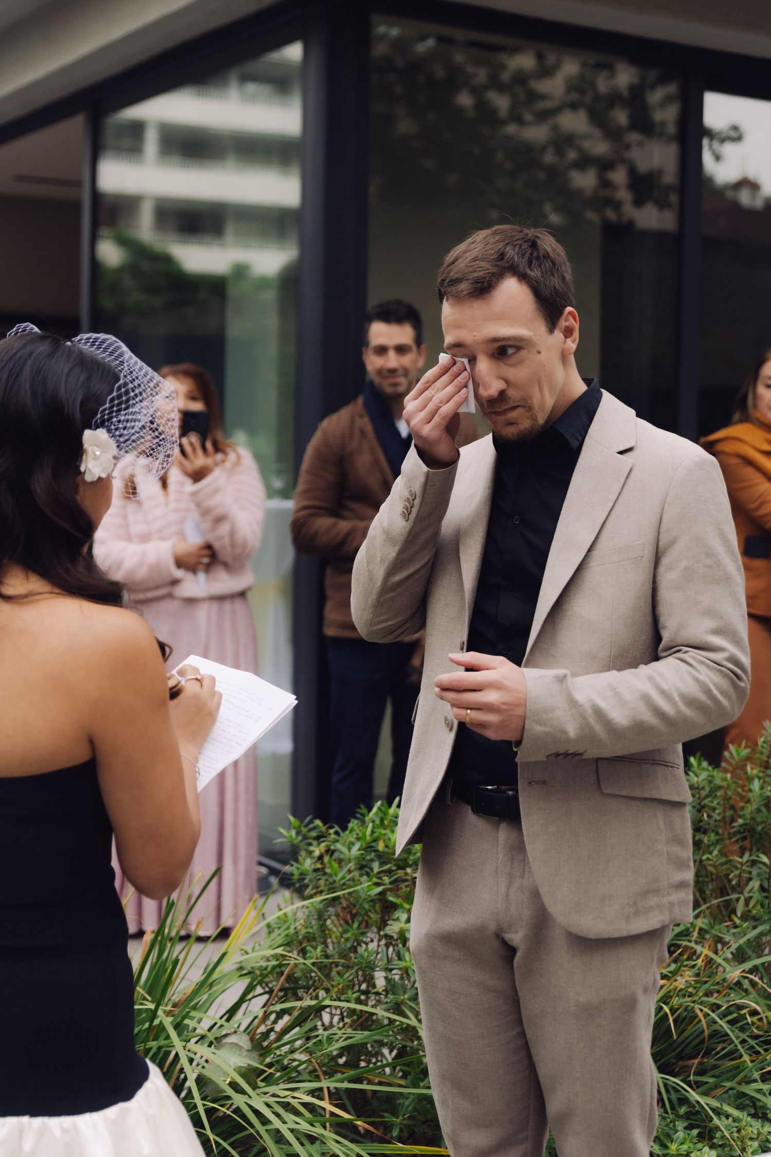 Emotional groom wiping tears while bride reads vows at outdoor wedding ceremony.