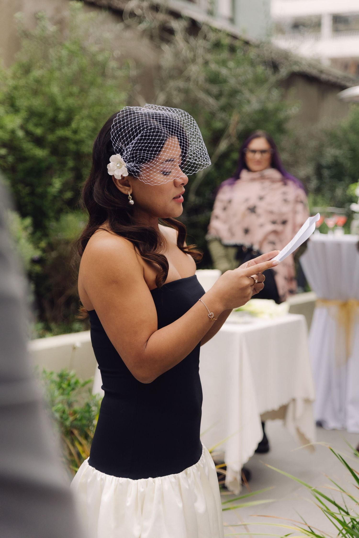 Bride in black dress and birdcage veil reading handwritten vows at outdoor wedding.