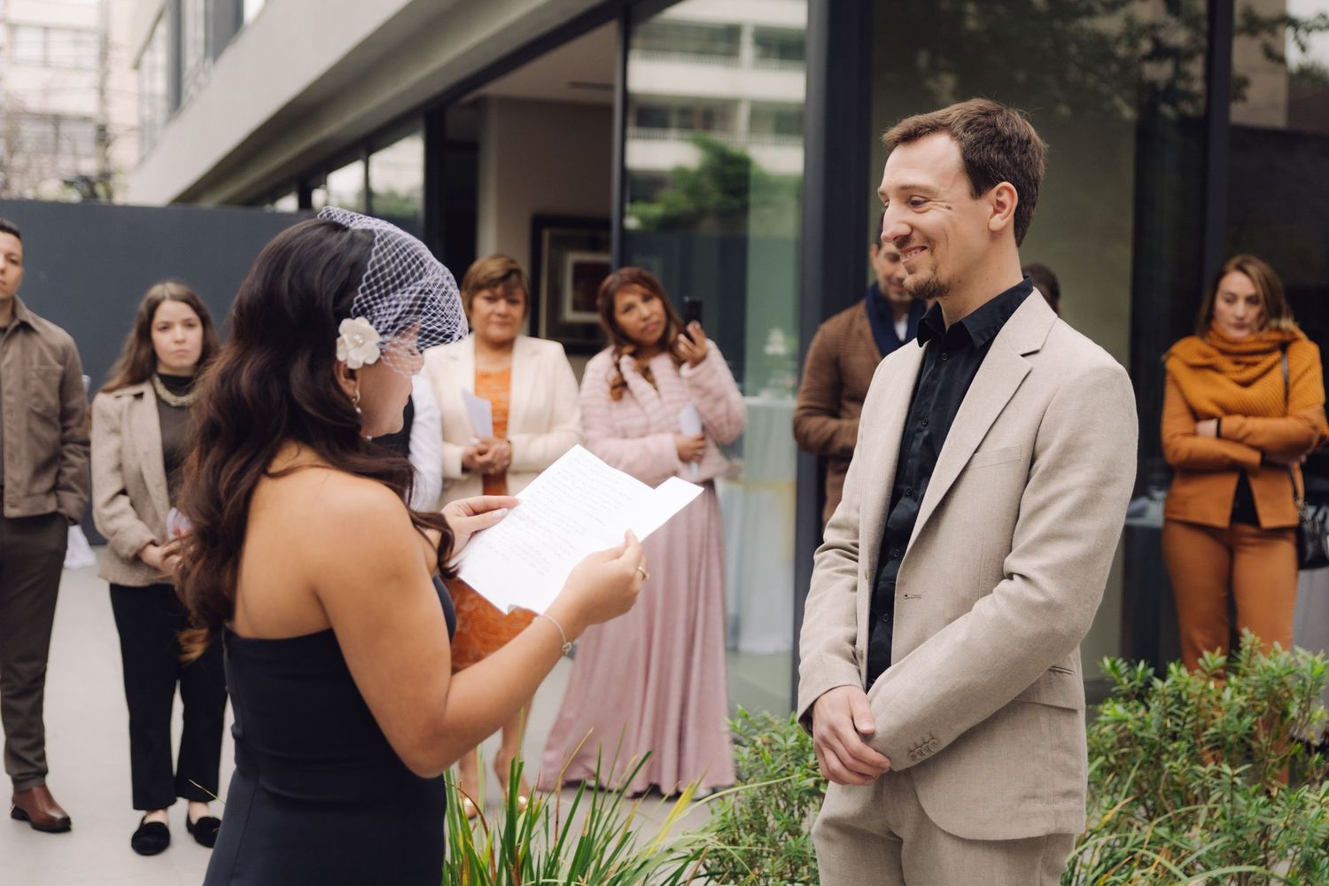 Bride reading wedding vows to groom at intimate outdoor ceremony with guests watching.