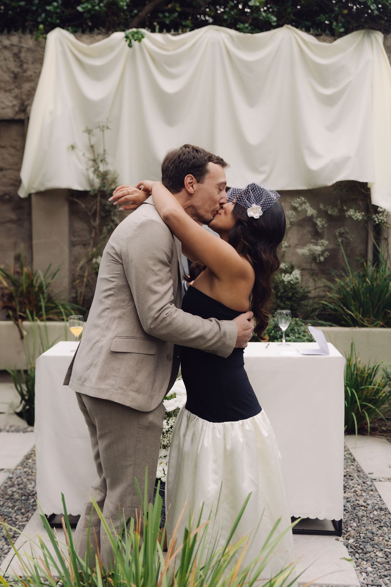 Newlyweds share a passionate kiss during intimate outdoor garden wedding ceremony with white drape backdrop.