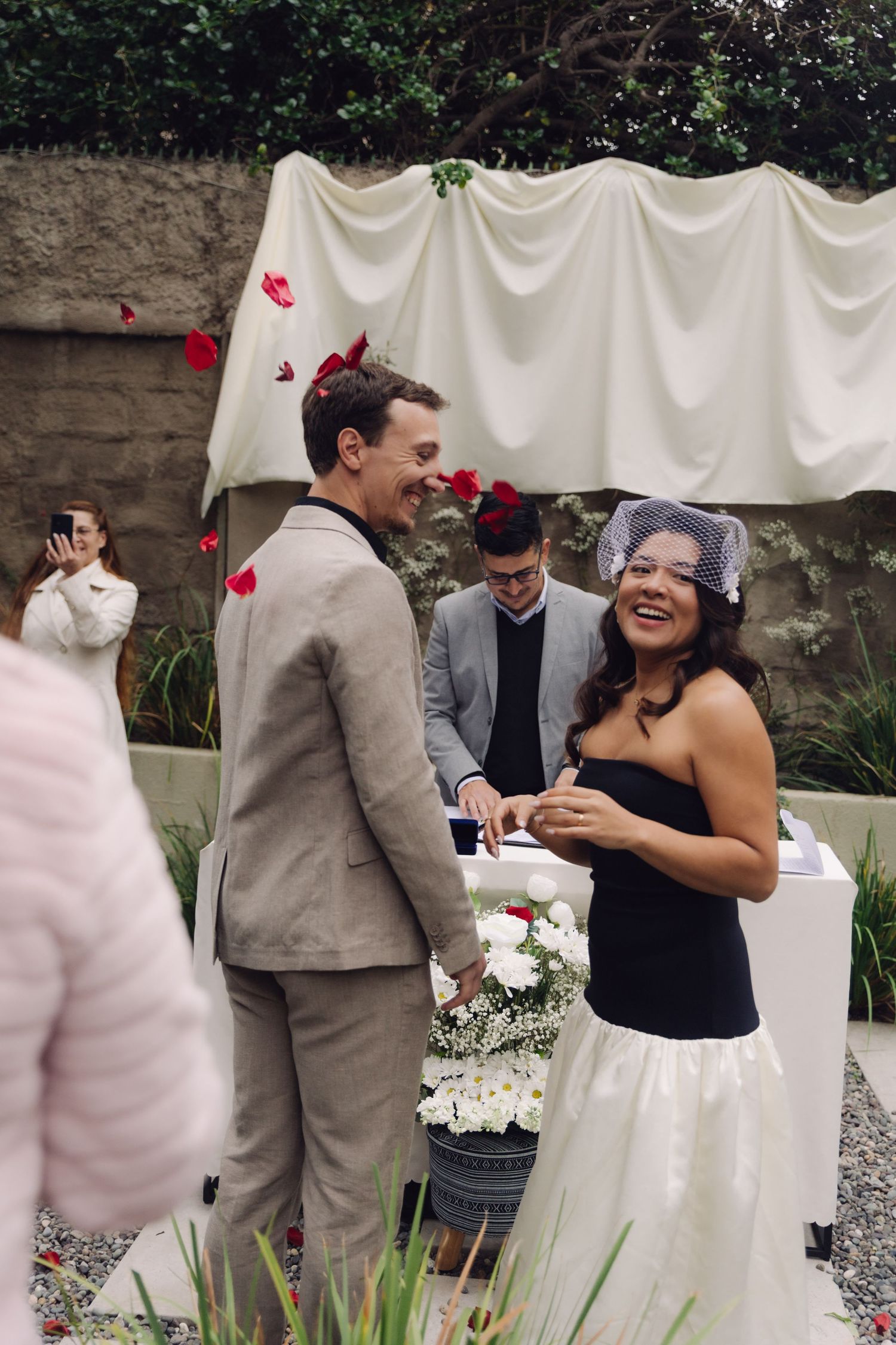 Bride and groom exchange rings at outdoor wedding ceremony with rose petals falling and officiant behind.