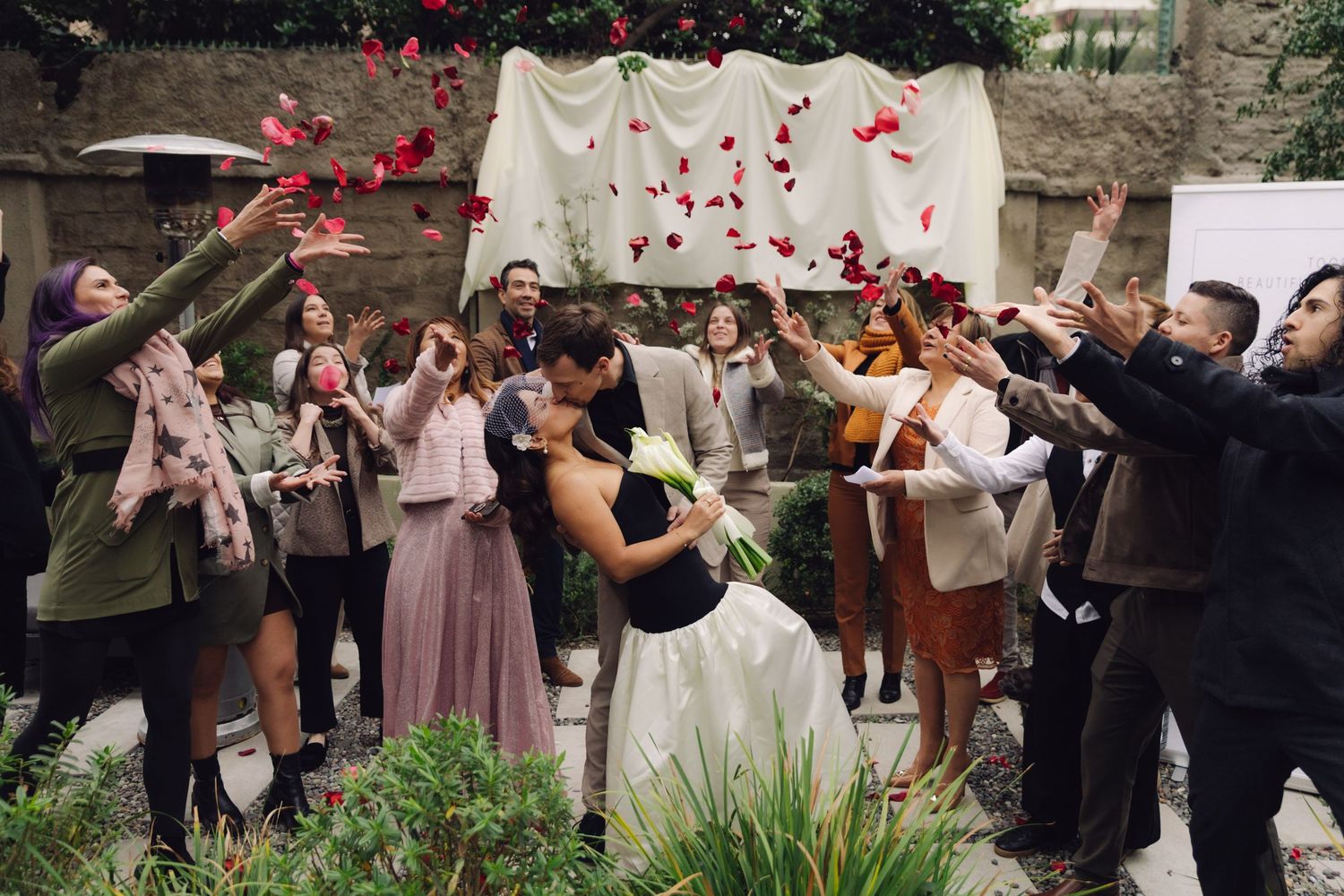 Wedding guests toss red rose petals as bride and groom kiss in joyful outdoor ceremony celebration.