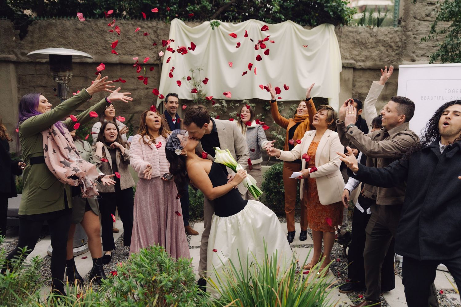 Guests shower newlyweds with red rose petals during romantic outdoor wedding ceremony kiss celebration.