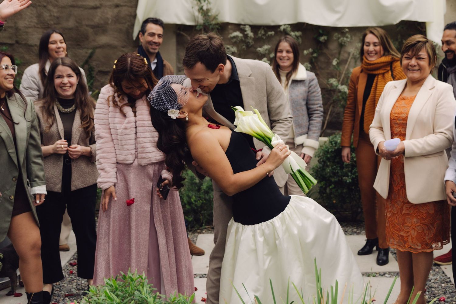 Groom dips bride for a romantic kiss surrounded by smiling wedding guests at outdoor ceremony.