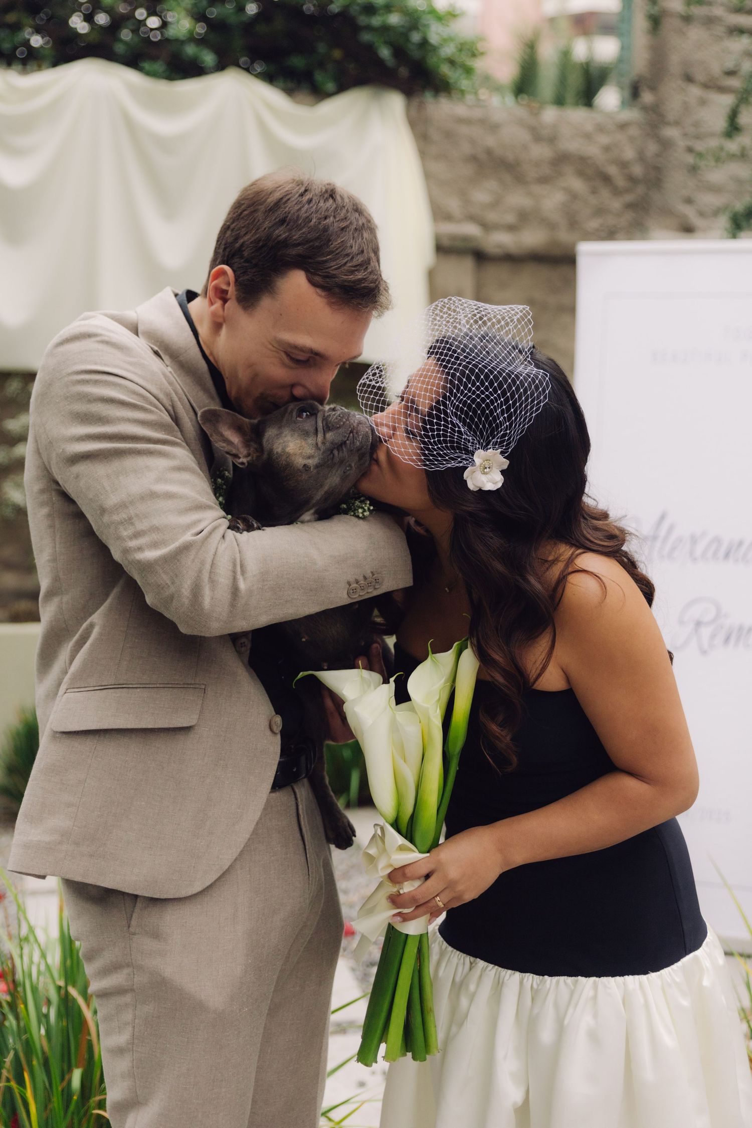 Bride and groom lovingly kiss their dog together at outdoor wedding ceremony holding calla lily bouquet.