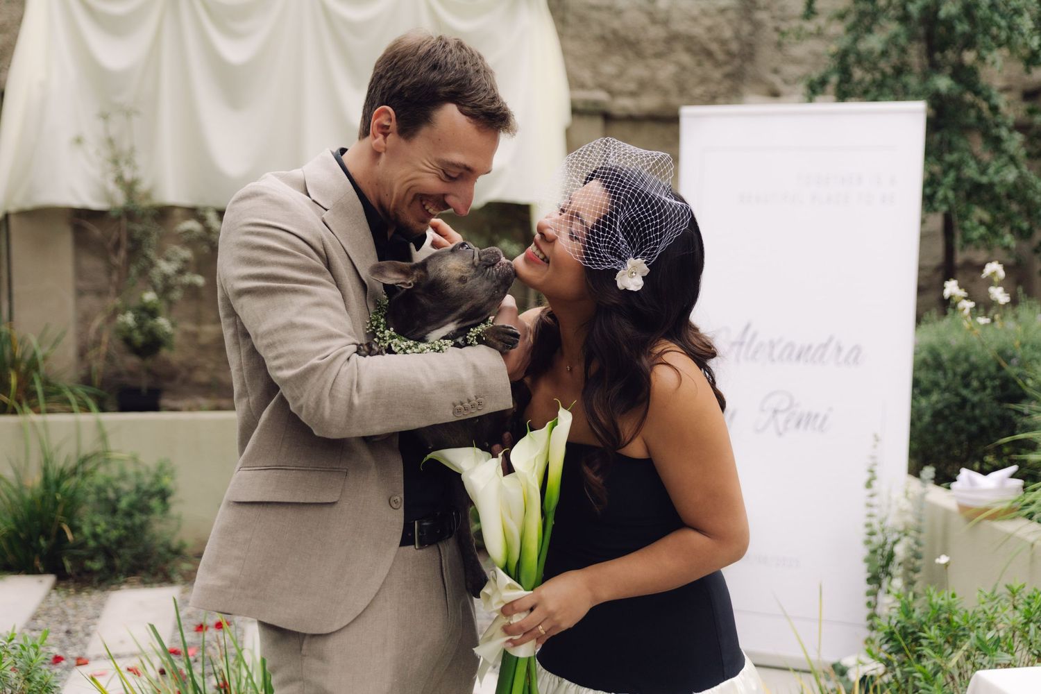 Happy newlyweds share a joyful moment with their dog at outdoor garden wedding ceremony.