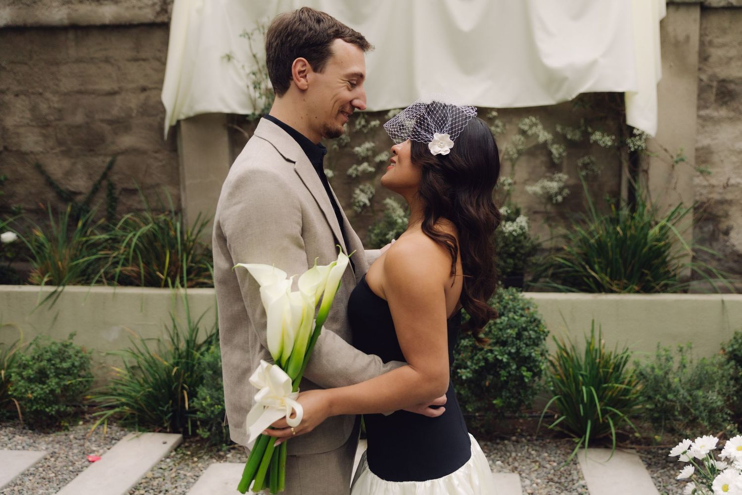 Bride and groom share a tender romantic gaze holding calla lilies at outdoor garden wedding.