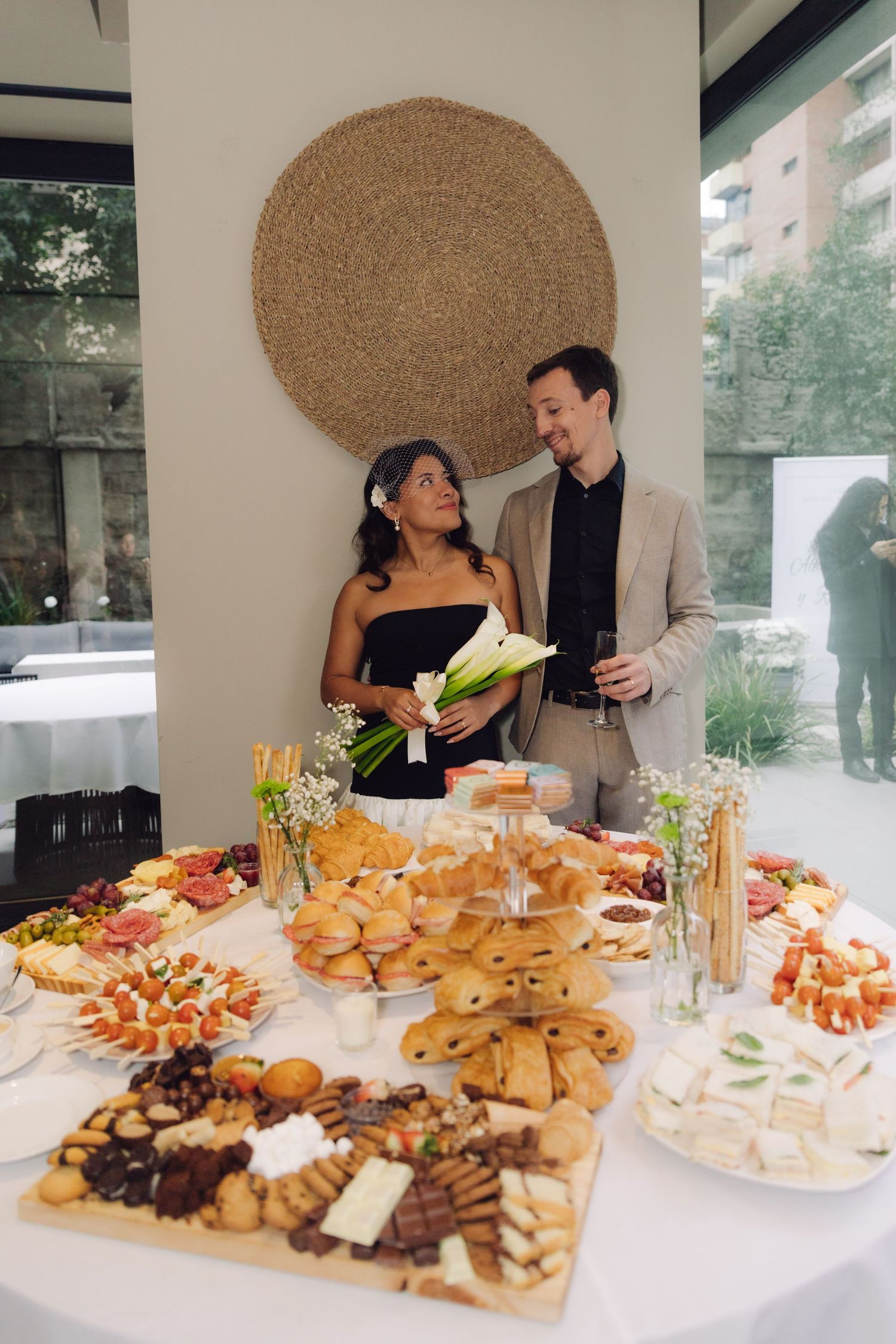 Newlywed couple standing beside elegant wedding reception grazing table with assorted foods.