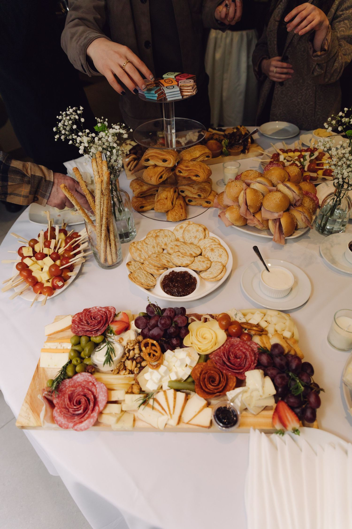 Guests reaching for food at wedding reception grazing table with charcuterie and pastries.