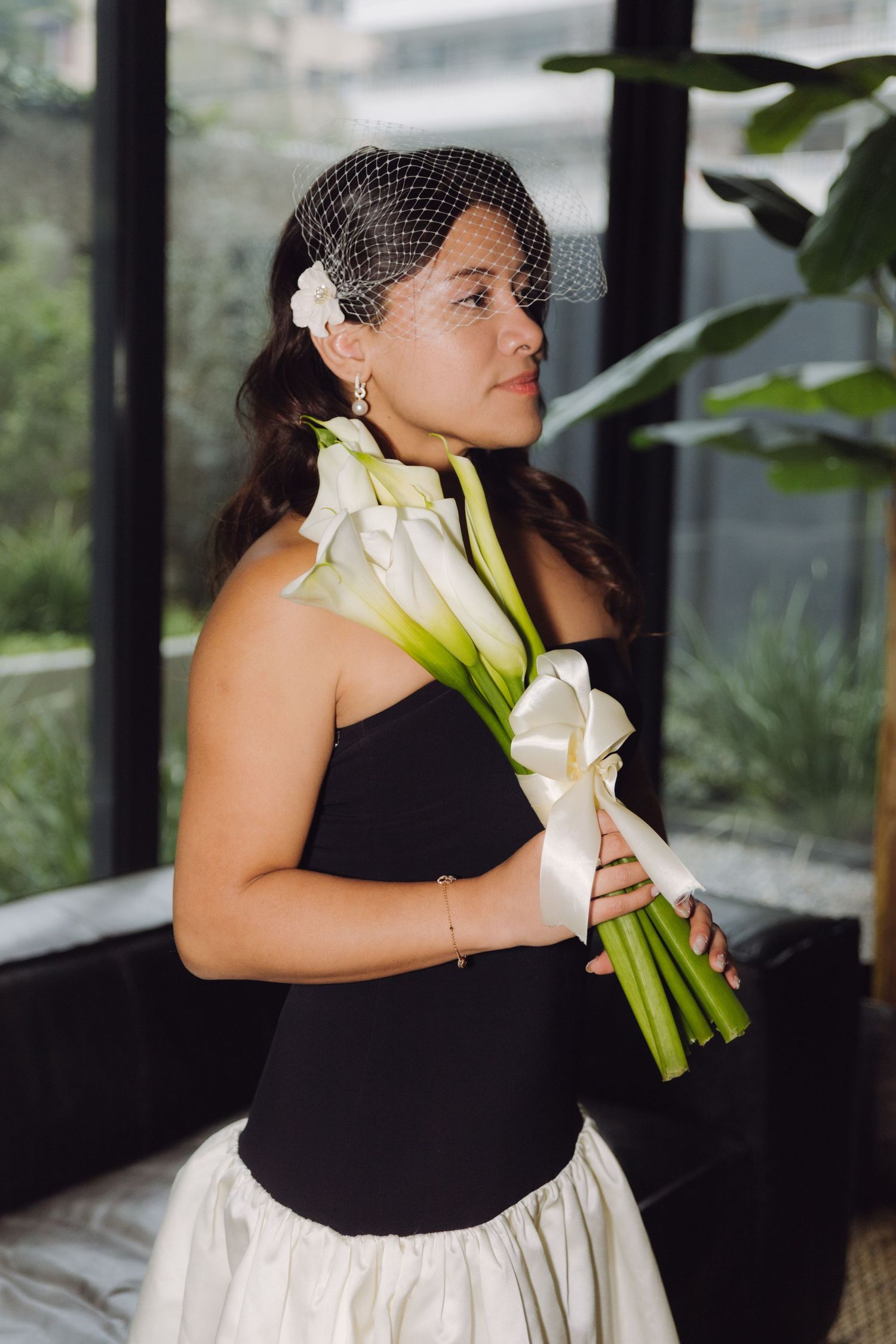 Bride in black strapless gown with white skirt holds calla lily bouquet wearing birdcage veil near window.