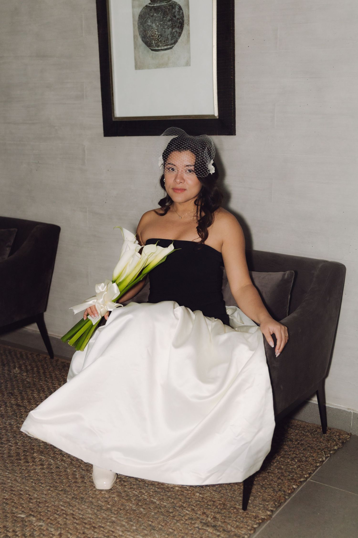 Elegant bride in black and white wedding dress sits on gray sofa holding white calla lily bouquet.