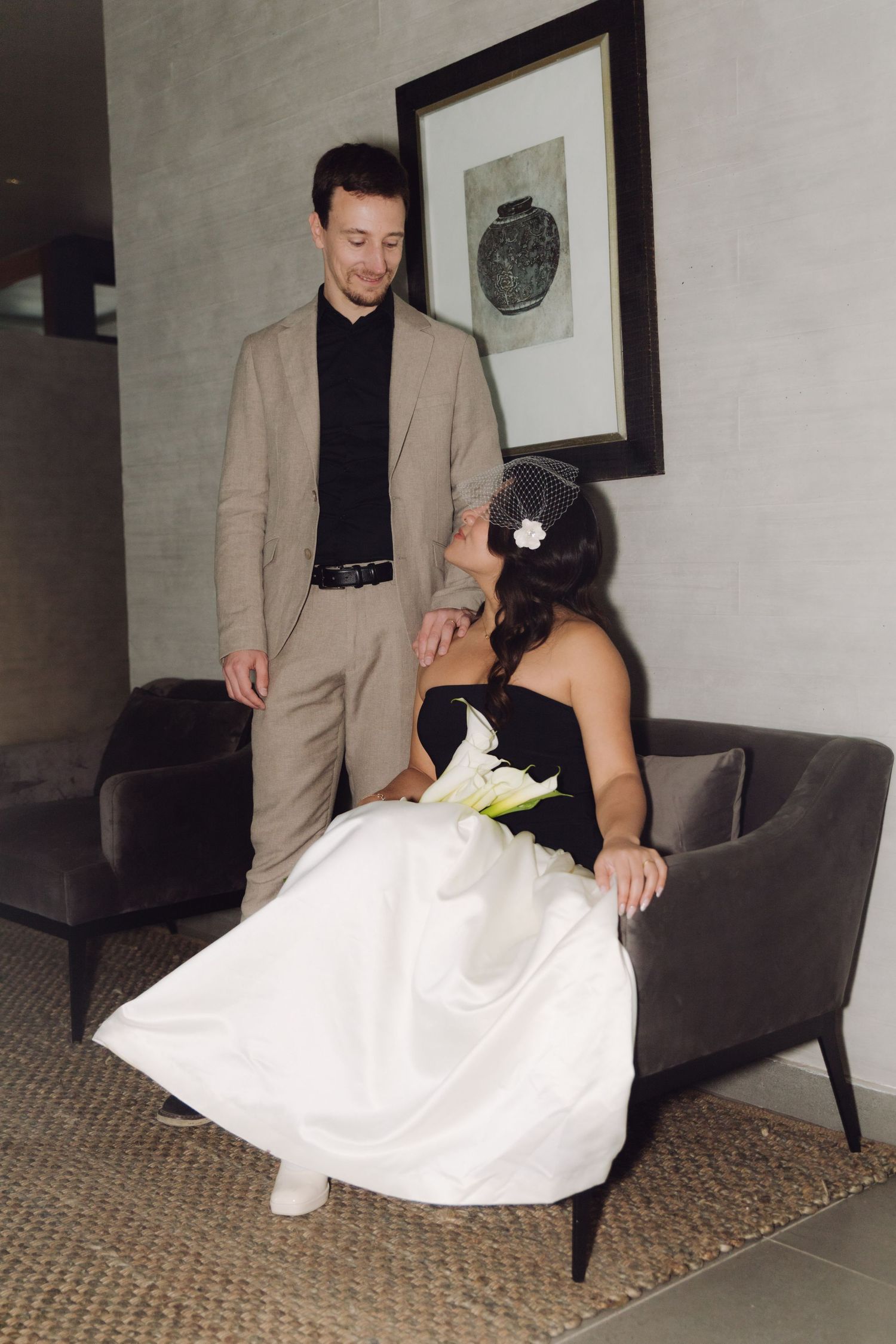 Groom in tan suit stands beside seated bride in black and white wedding dress in modern interior.