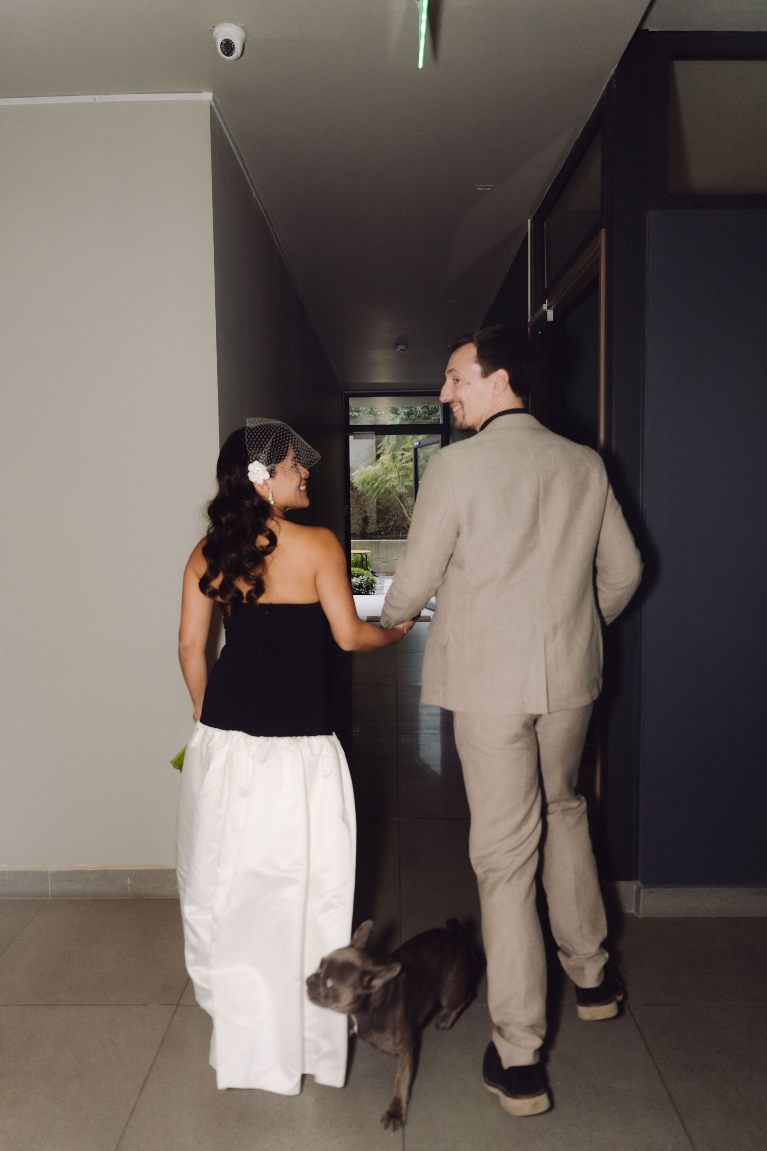 Bride and groom walk hand in hand down a modern hallway with their gray dog.