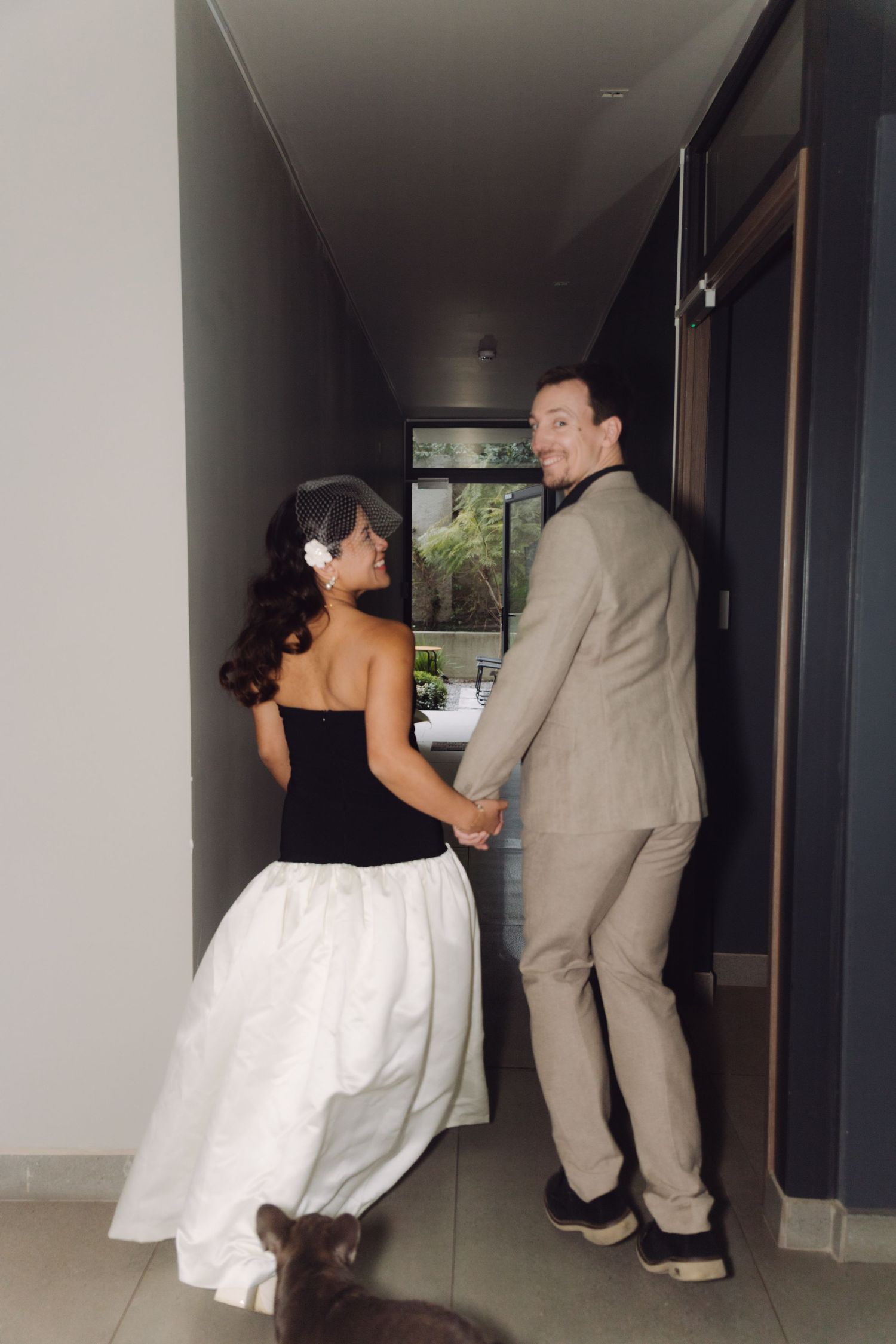 Bride and groom hold hands in a hallway doorway as their dog watches from below.