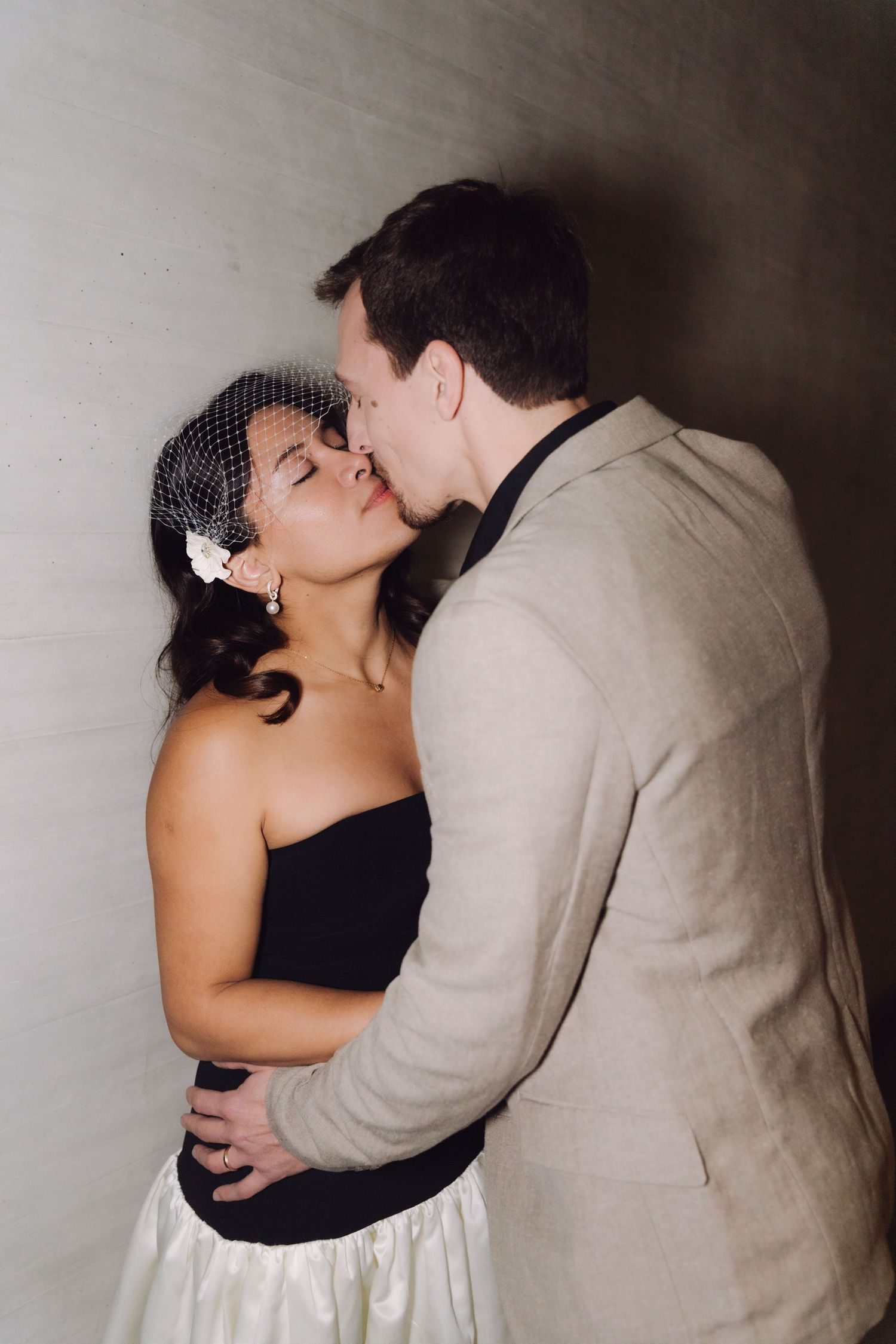Groom tenderly kisses bride wearing birdcage veil as they embrace against a gray wall.