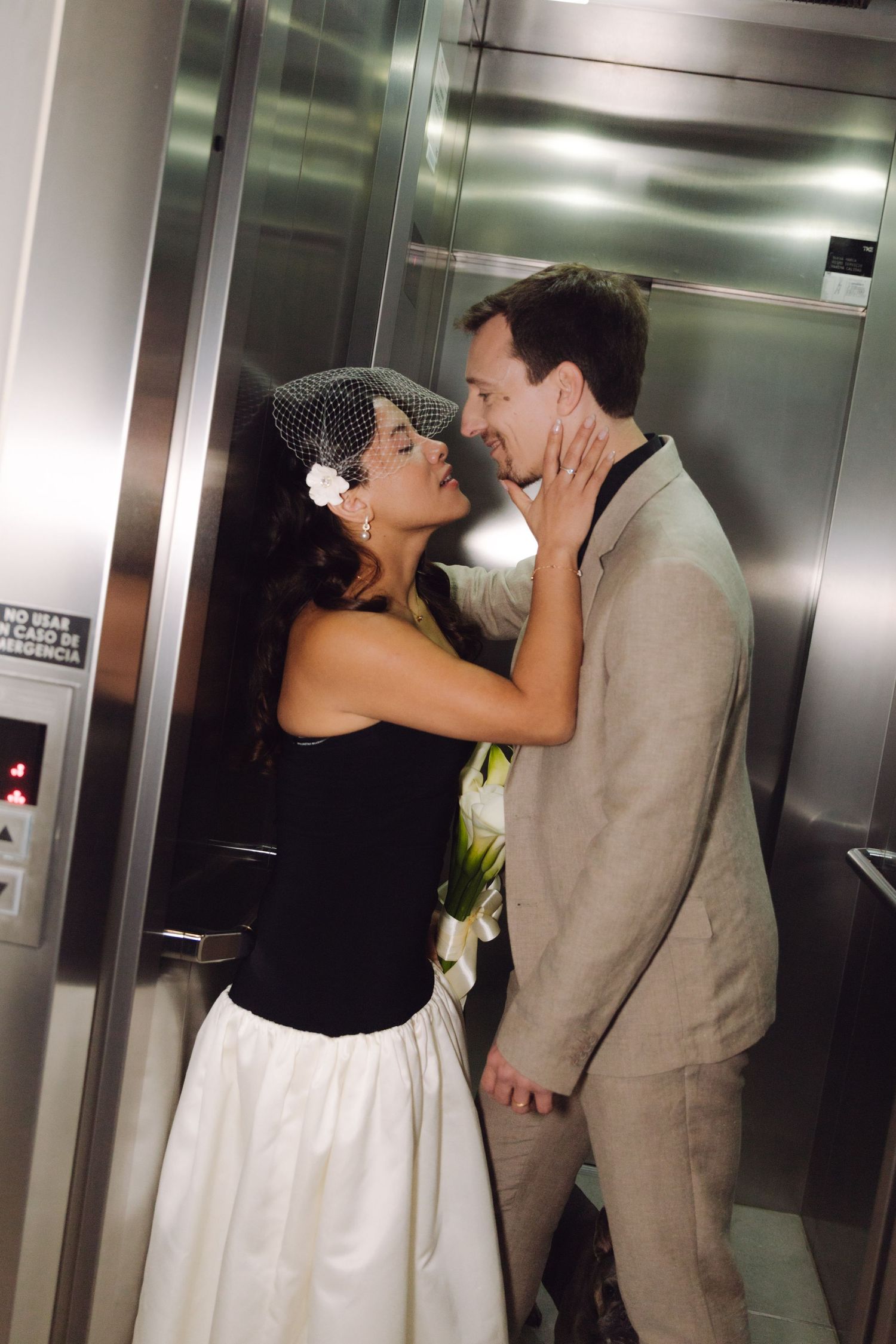 Bride in birdcage veil and groom share a romantic embrace inside a silver elevator.